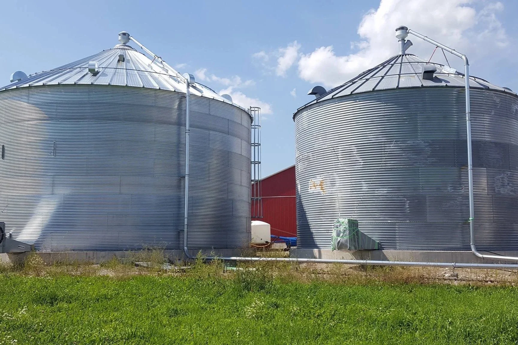 Two large shiny metal grain silos with a living roof, connected by pipes, on a farm with a red barn in the background and a well-maintained grassy area in the foreground.