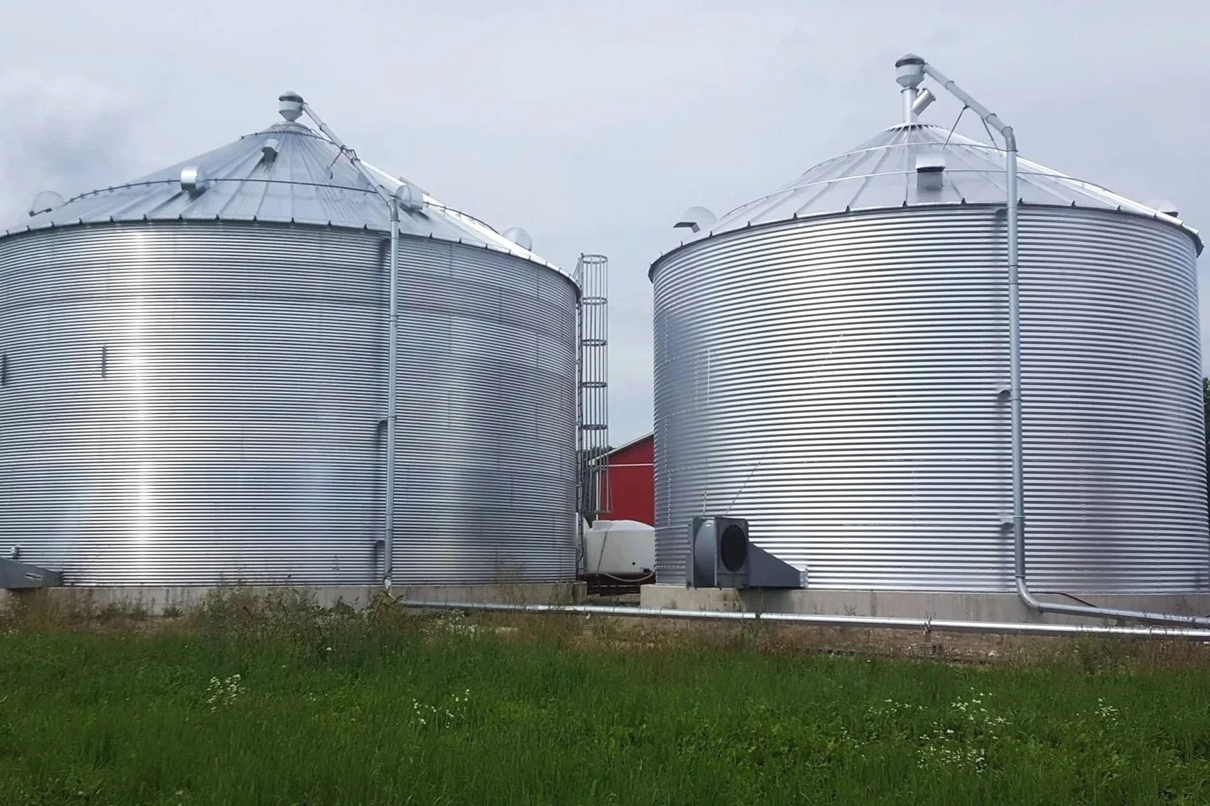 Two large silver grain silos with metallic roofs and attached pipes in a farm setting with green grass and cloudy sky.