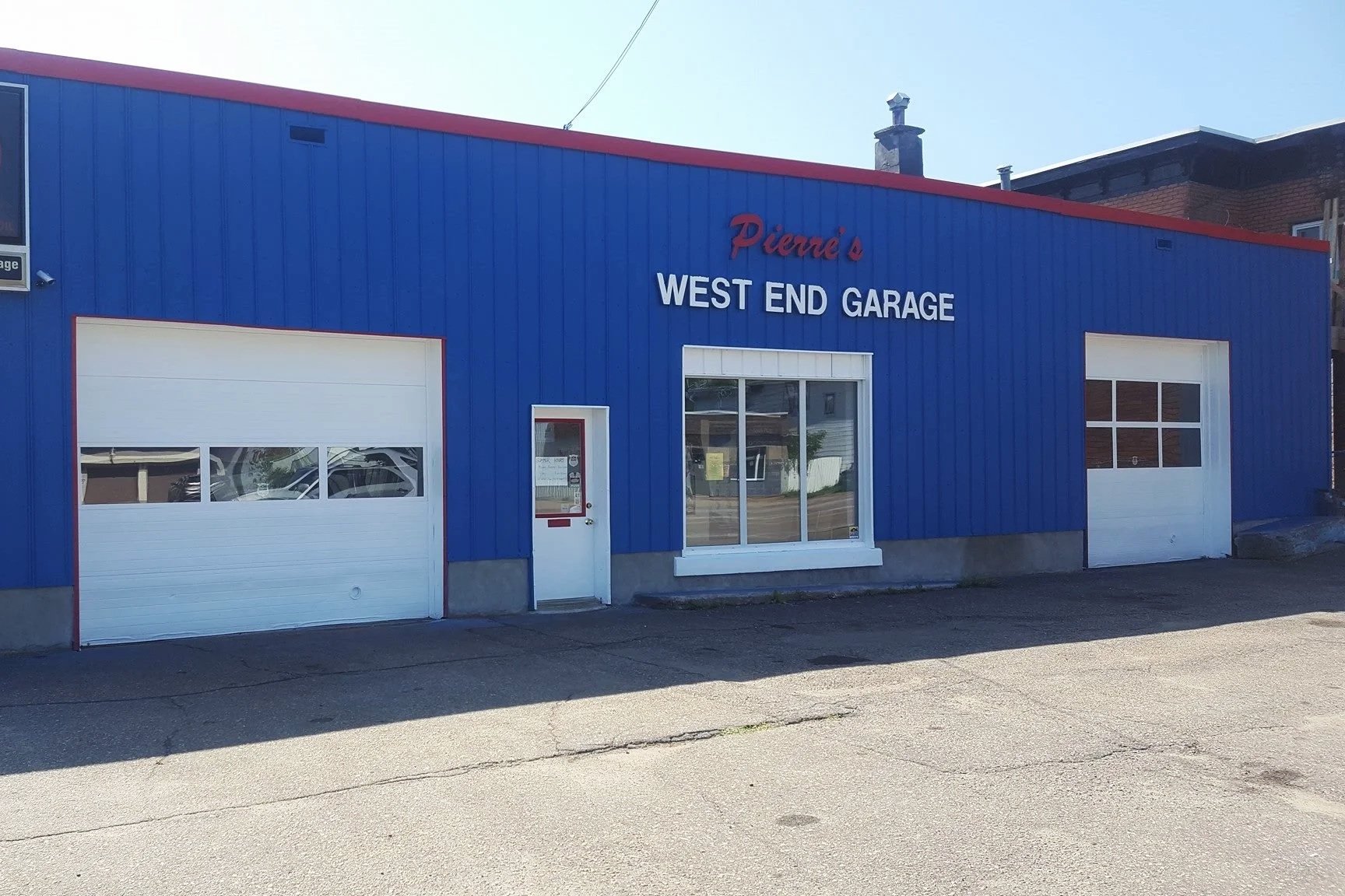 The image shows a blue garage building with two white garage doors and a small entrance door in the middle. The building has red accents and signage that reads "Pierre's WEST END GARAGE." There is a large window next to the entrance door.