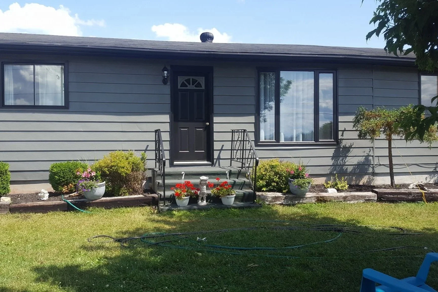 Front of a gray house with black door, windows, and small front garden with potted plants, flowers, and a lawn