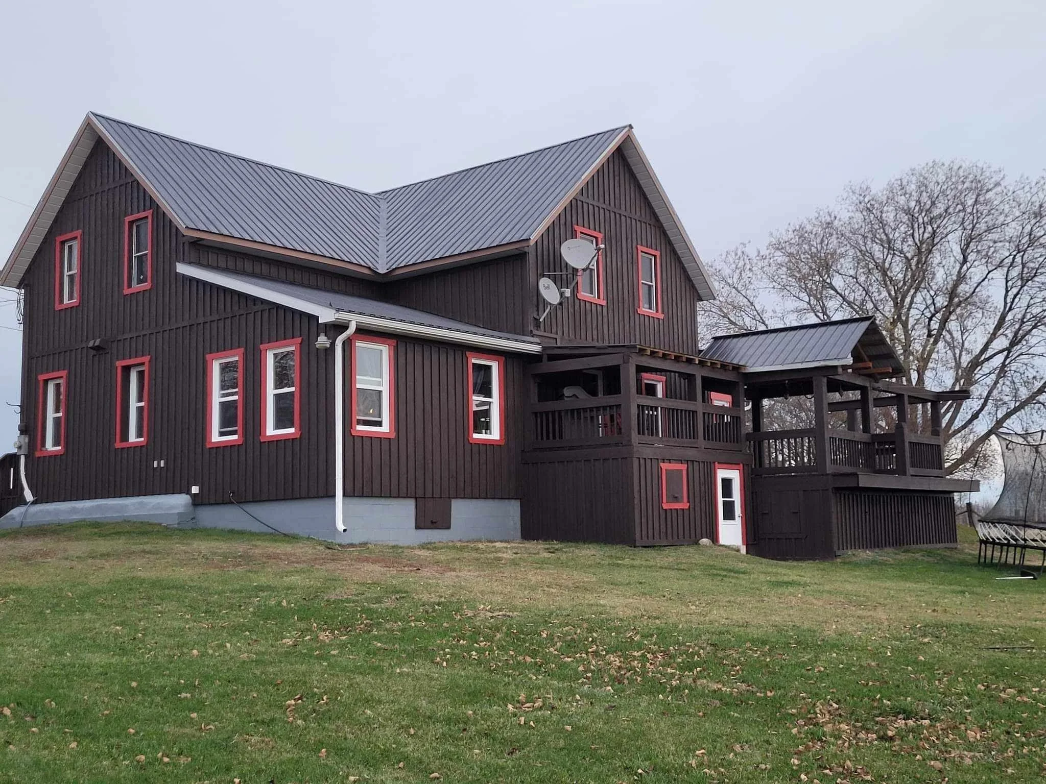 A dark brown wooden house with a metal roof and red trim around the windows, situated in a lawn with grass and fallen leaves, with leafless trees in the background.