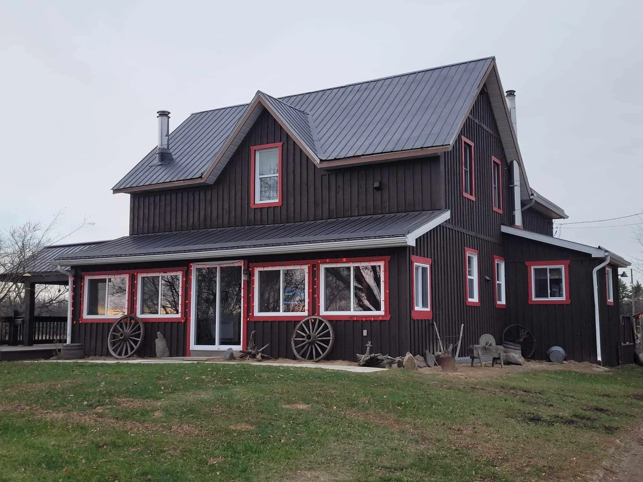 A two-story house with black wooden siding and red window frames. The house has a steep metal roof with chimneys and is decorated with red string lights. Several old wagon wheels are used as decoration at the front.