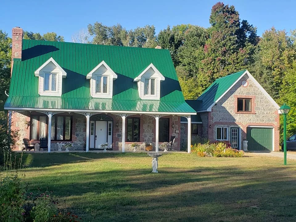 Stone house with green metal roof, dormer windows, front porch, and attached garage, surrounded by trees and a well-kept lawn.