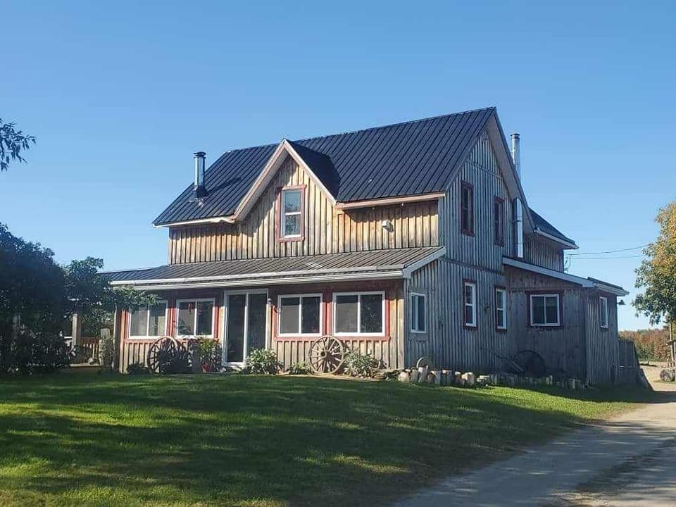 A rustic two-story house with a metal roof, wooden siding, and multiple windows, surrounded by a grassy lawn and trees.
