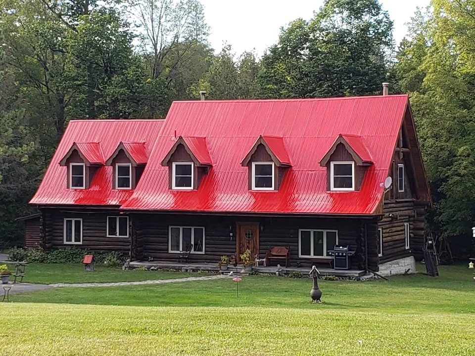 A large log cabin with a red metal roof and four dormer windows, surrounded by green grass and trees.