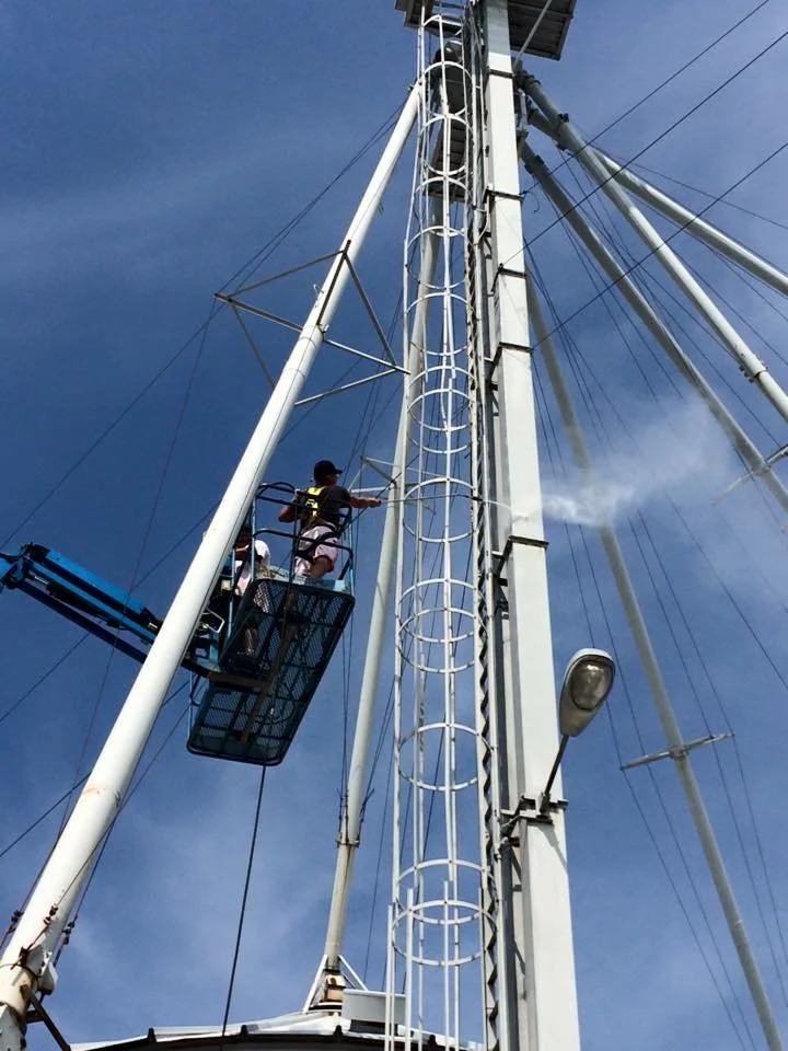 A person standing in a cherry picker platform washing a tall communication tower against a blue sky.