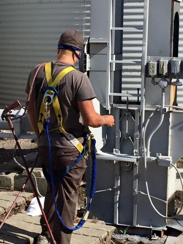 Worker inspecting electrical box on a utility pole, wearing safety harness, helmet, and gloves.