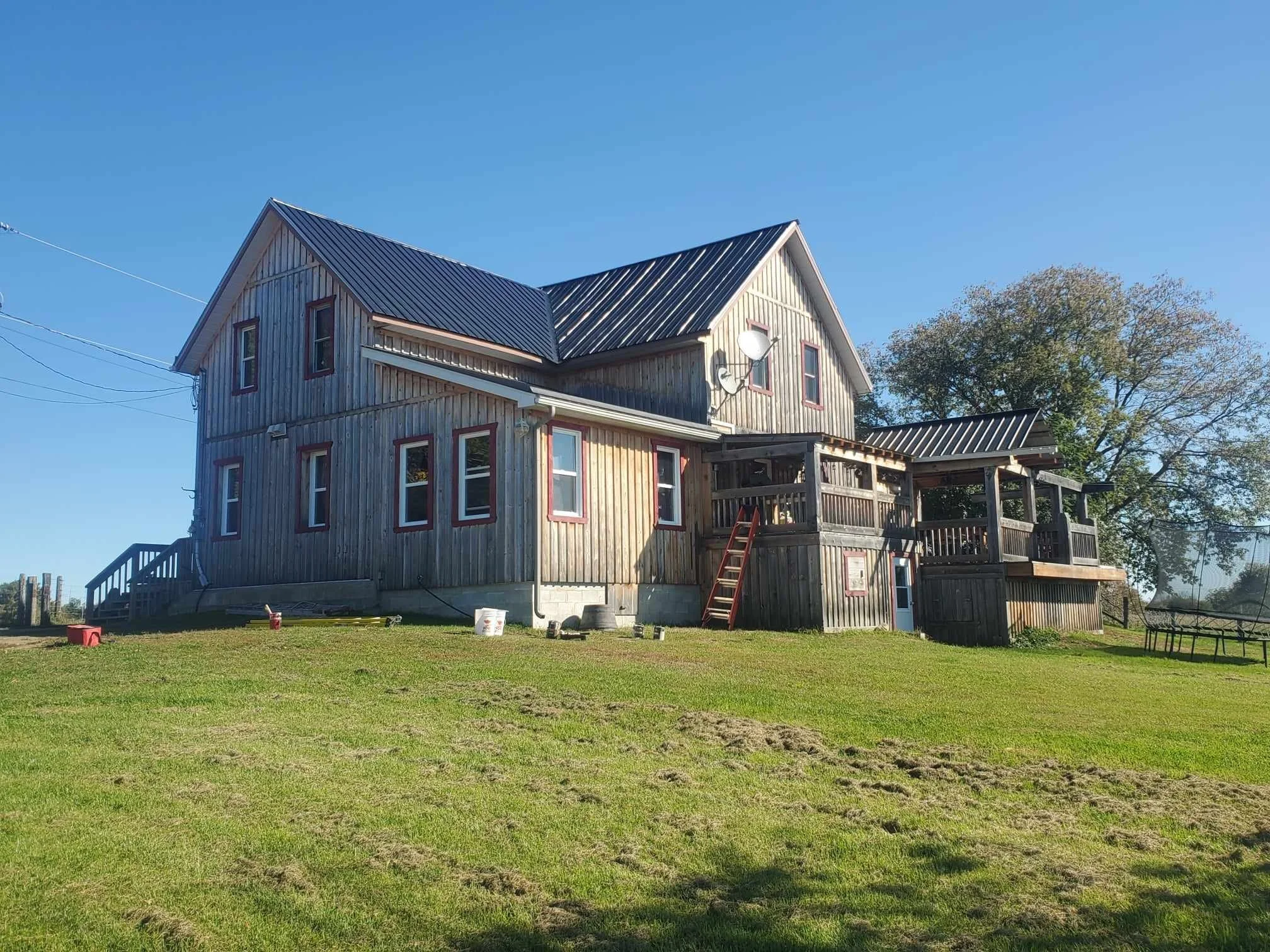 A two-story wooden house with a metal roof, multiple windows, and a small deck with stairs, set in a grassy yard with a tree and blue sky in the background.