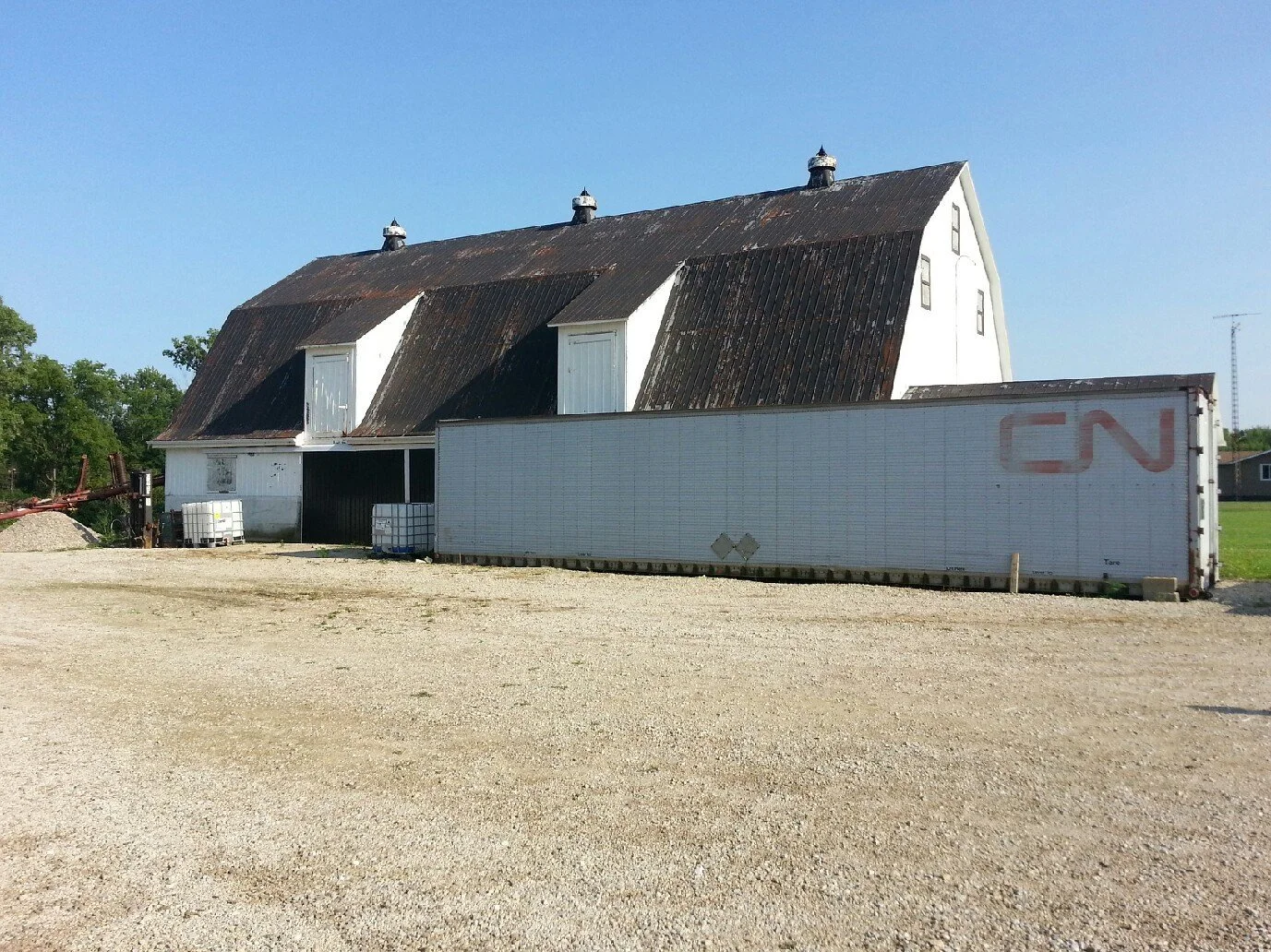 Old white barn with a rusty roof, a large shipping container in front, and a few water tanks beside it, under a clear blue sky.