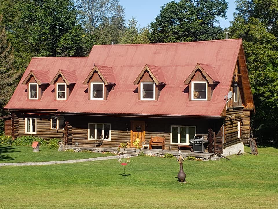 A two-story log cabin with a red metal roof, four dormer windows, a wooden front door, and a porch with outdoor furniture and a barbecue grill, surrounded by green grass and trees.