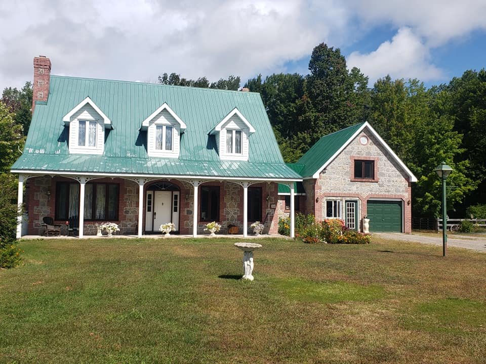 A large house with a green metal roof, stone exterior, and white trim. There are three dormer windows on the roof, and a front porch with columns. The house has a garage on the right side with a green door. The front yard features a grassy lawn, pott