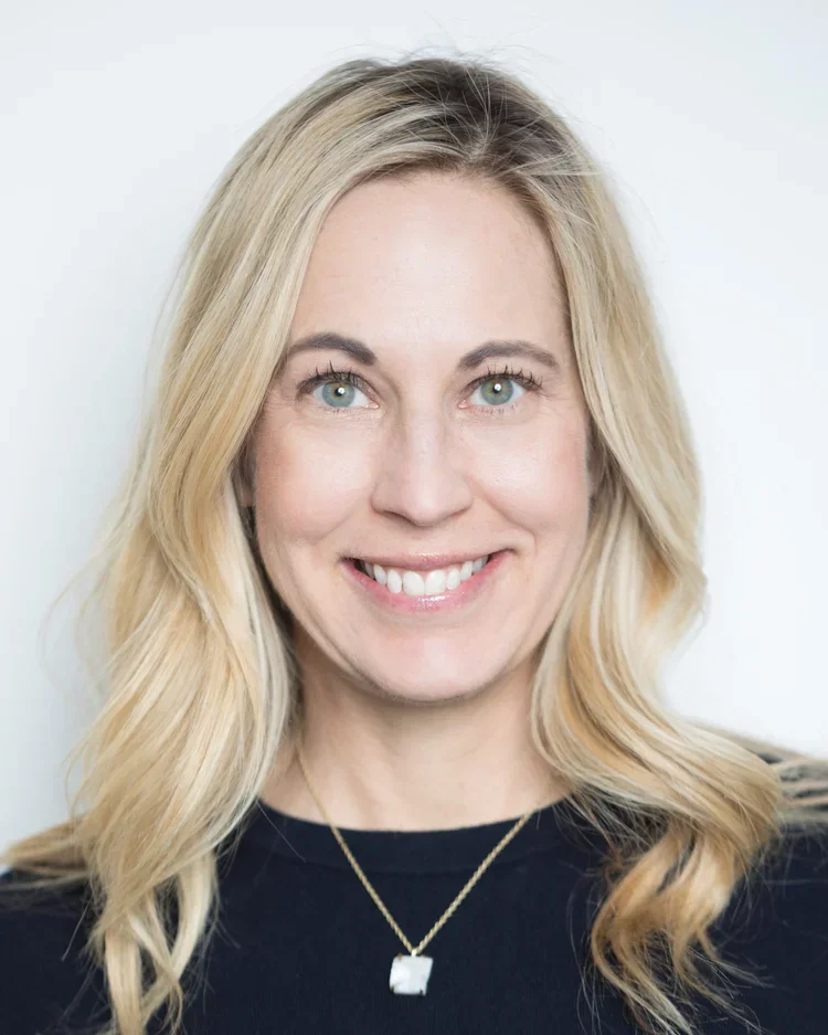 A smiling blonde woman with blue eyes, wearing a black top and a gold necklace with a small square pendant, against a plain white background.
