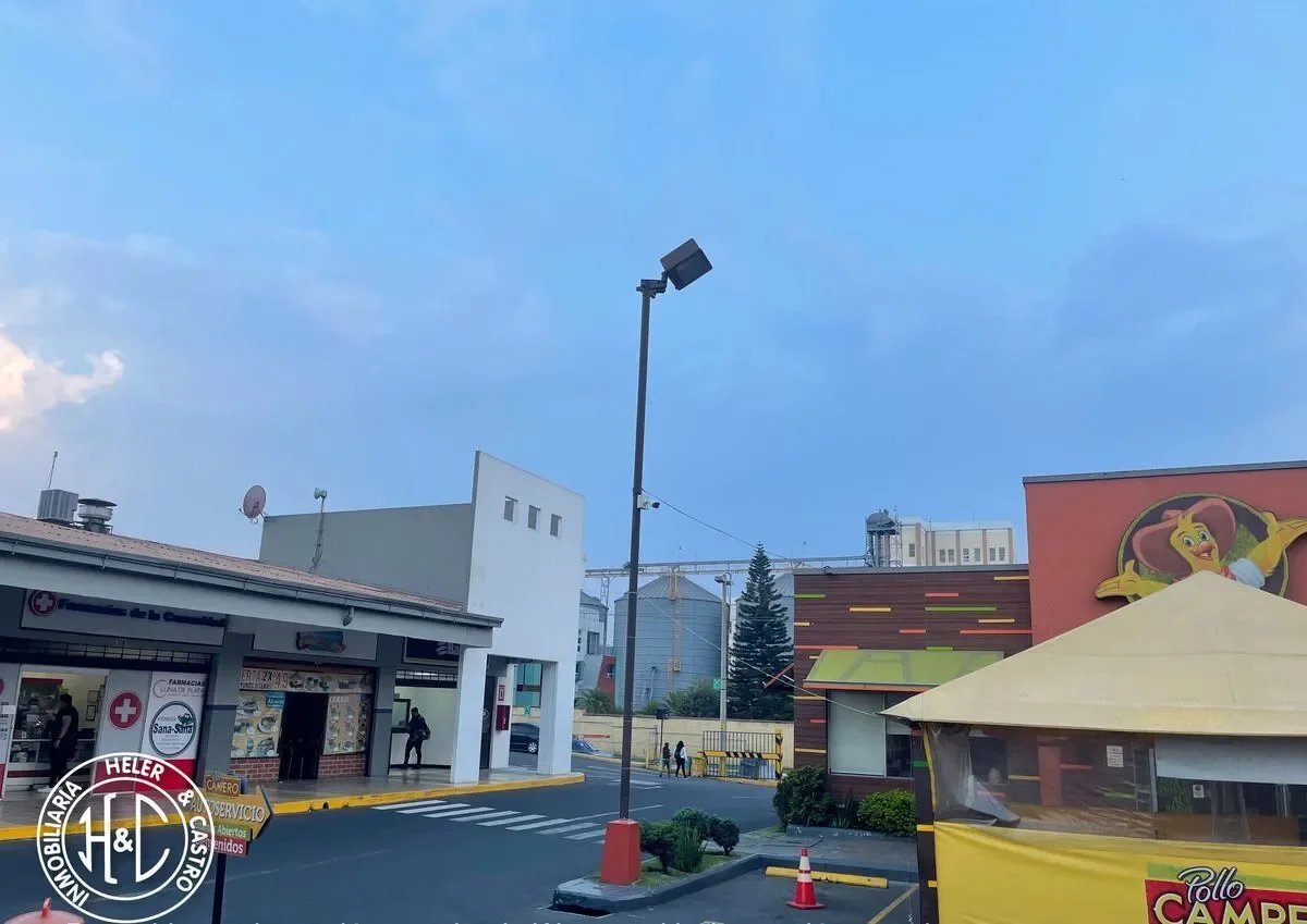 Vista de una zona comercial con una farmacia a la izquierda, una estructura de comida rápida en la esquina derecha, y un edificio decorado con una imagen de la caricatura de la selva. Hay una lámpara en el centro y varias personas en el fondo.