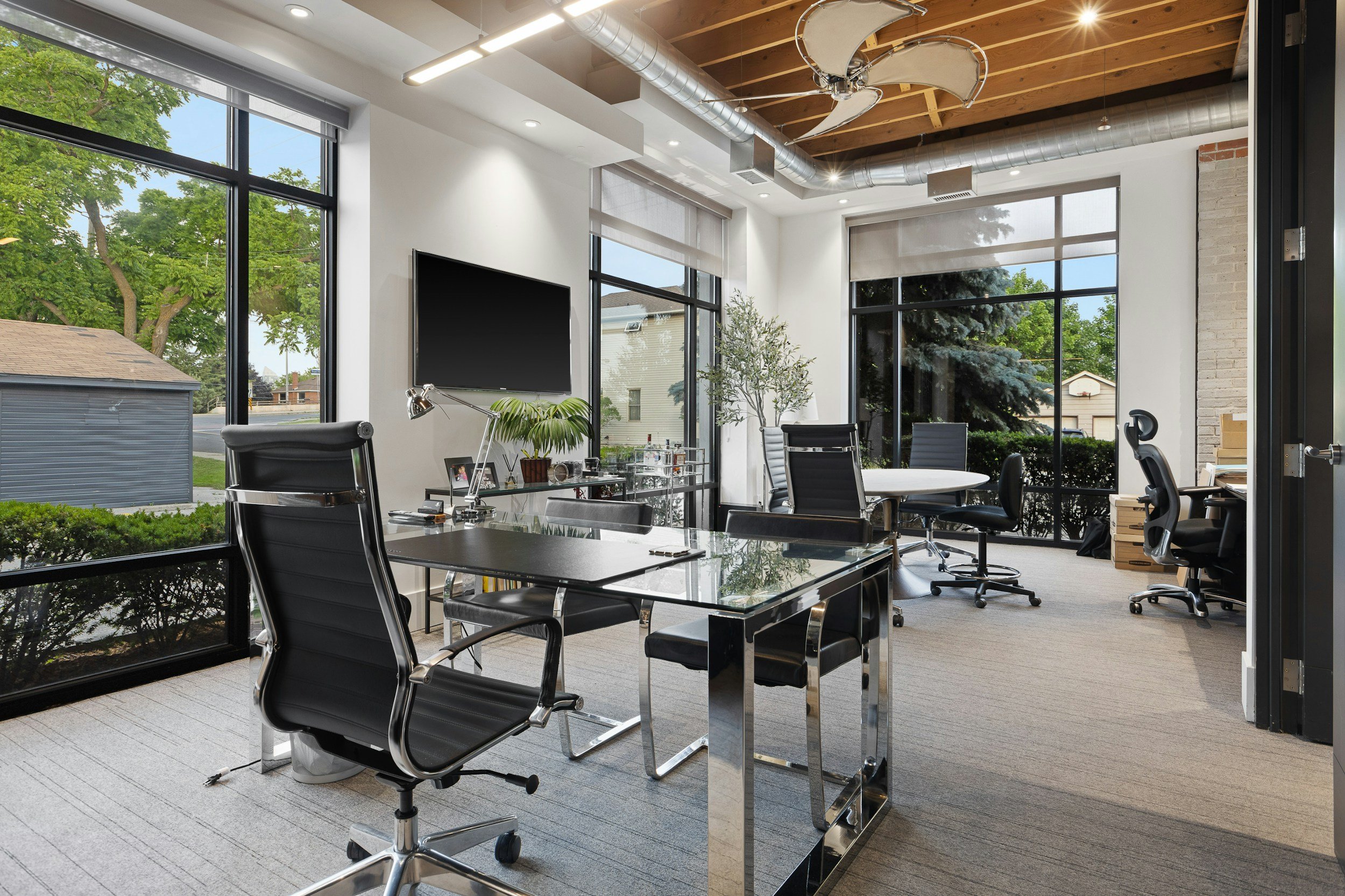 Modern office conference room with large floor-to-ceiling windows showing green trees and neighboring houses, furnished with black chairs, a glass table, and a wall-mounted television, with wood and brick accents and ceiling ducts.