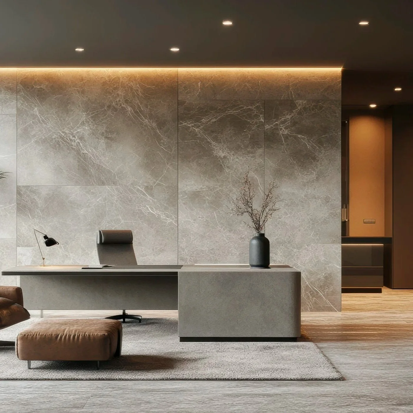 Modern office reception area with a gray desk, black office chair, brown ottoman, gray wall with marble texture, black vase with branches, and ceiling lights.