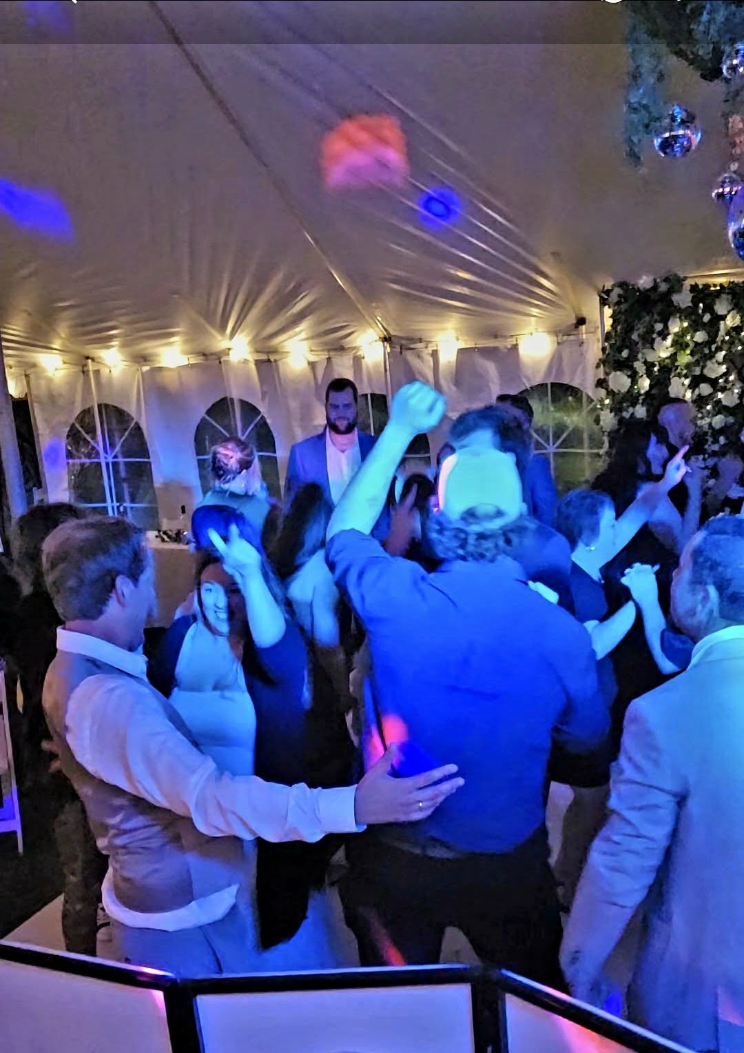People dancing and celebrating inside a decorated event tent with string lights and floral arrangements, under colorful lighting.