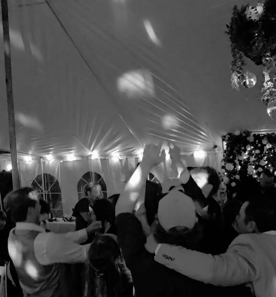 People dancing at a party or celebration inside a decorated tent with string lights and a Christmas tree.