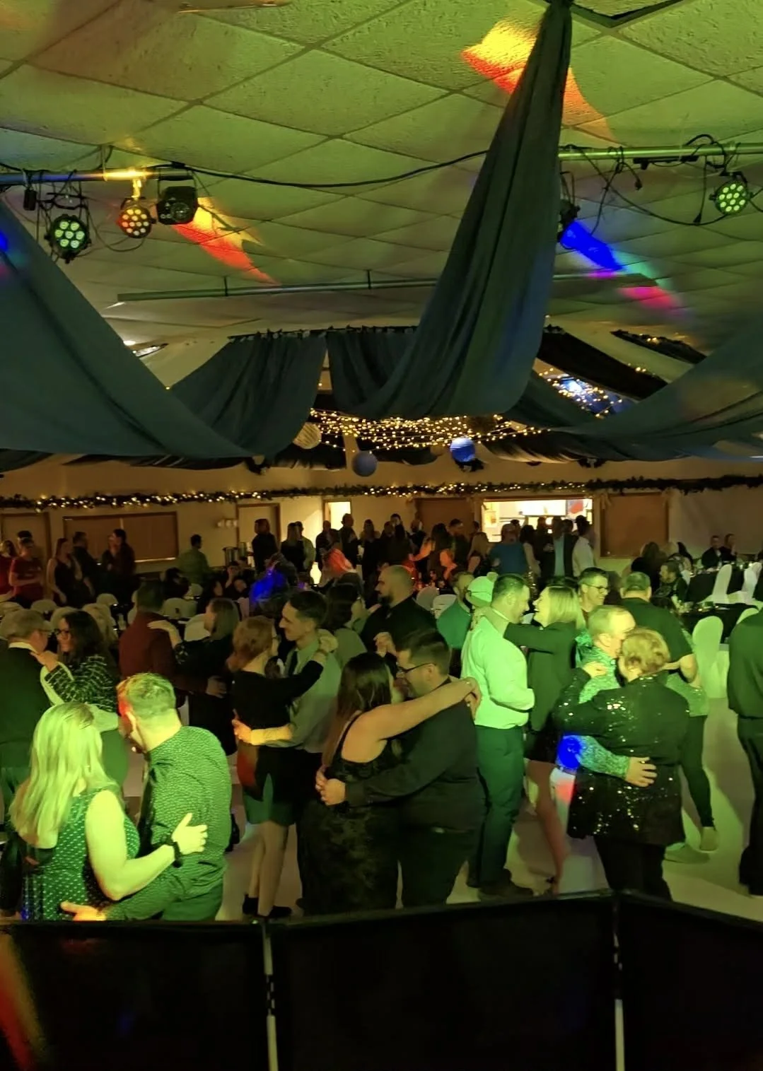 A crowded indoor dance party with people dancing and socializing. The ceiling has colorful lights and fabric drapery, with string lights and decorations.