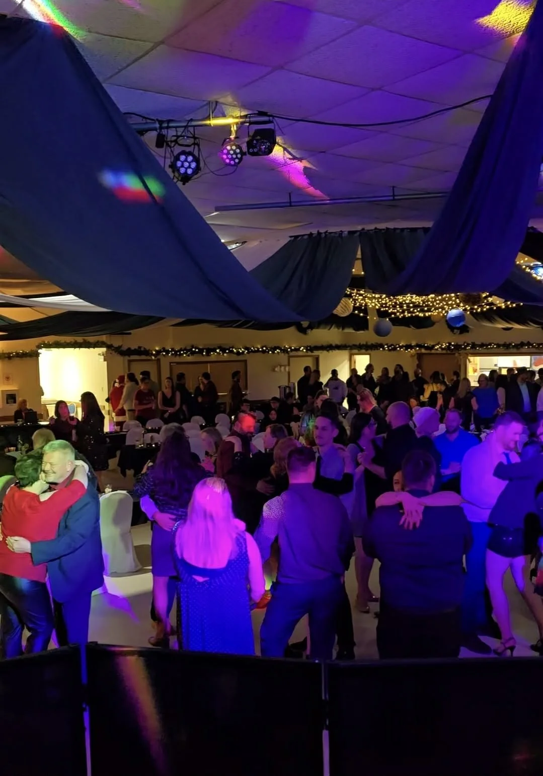 People dancing and socializing at a lively indoor party or celebration with festive decorations, draped fabric, string lights, and colorful lighting.