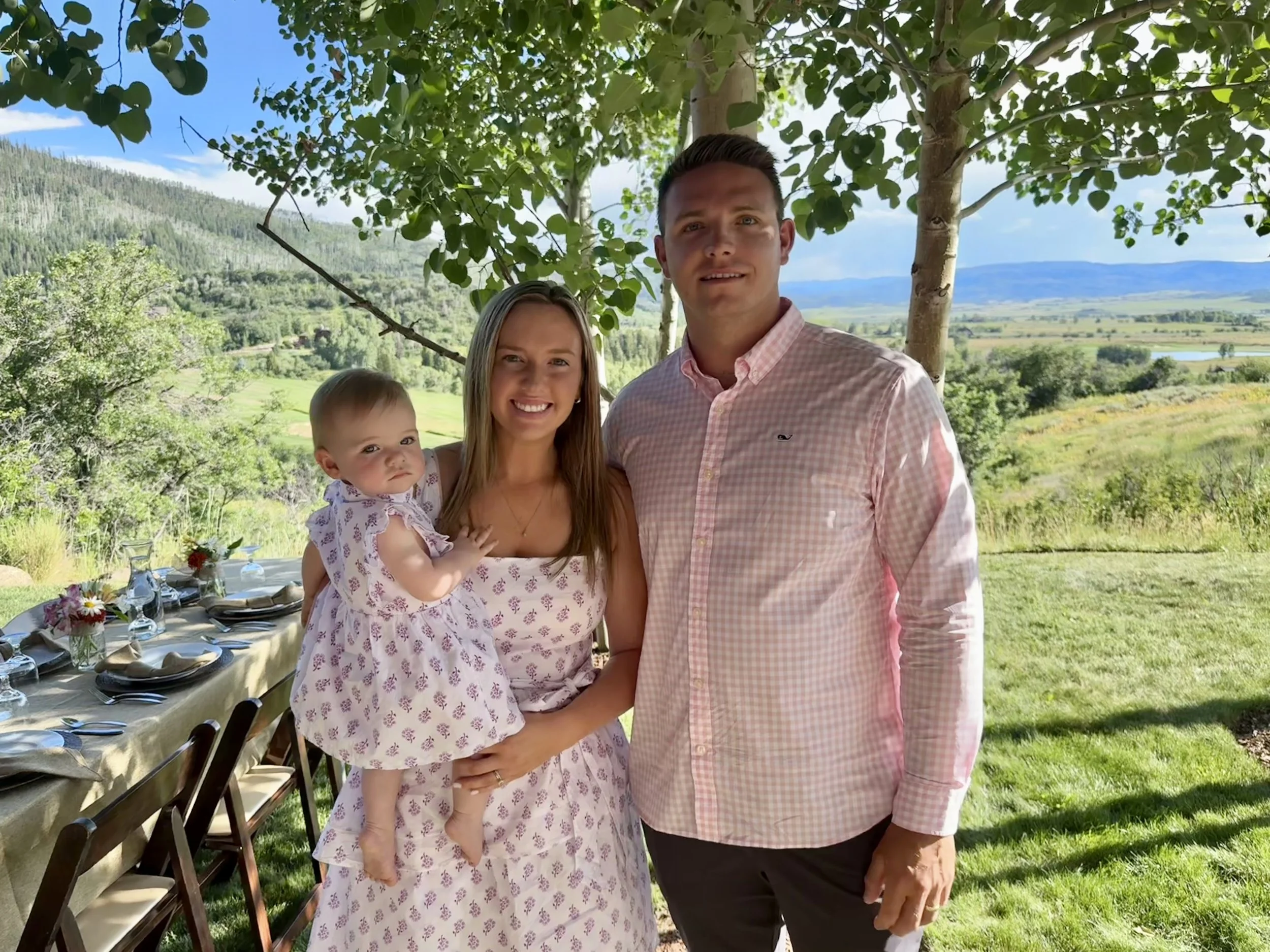 A family of three standing outdoors under a tree, with a scenic view of rolling hills, trees, and a blue sky in the background. The family includes a woman holding a young girl, and a man beside them.