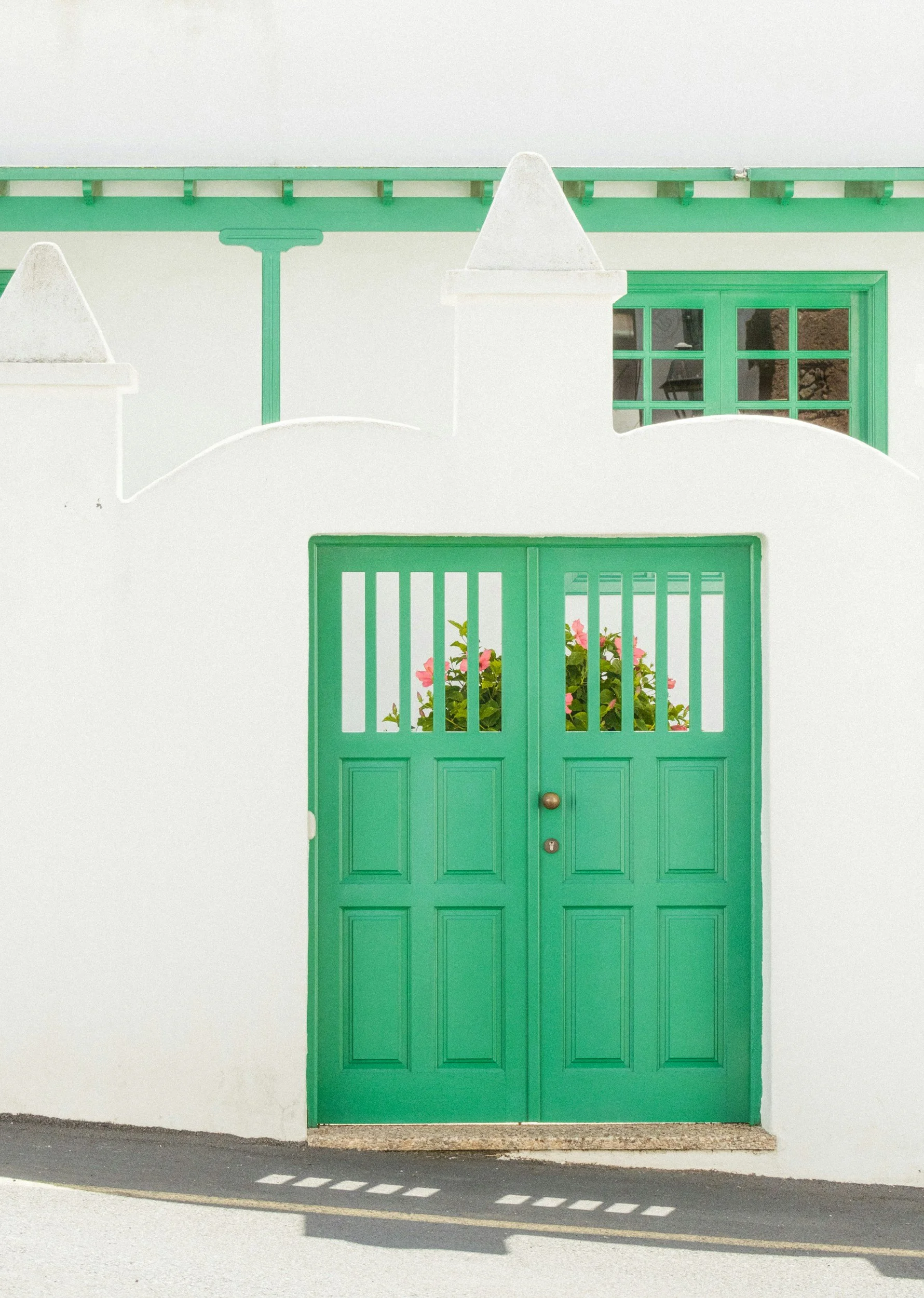 Green double door with window panels and a flowerpot with pink flowers, set against a white wall with decorative white posts.