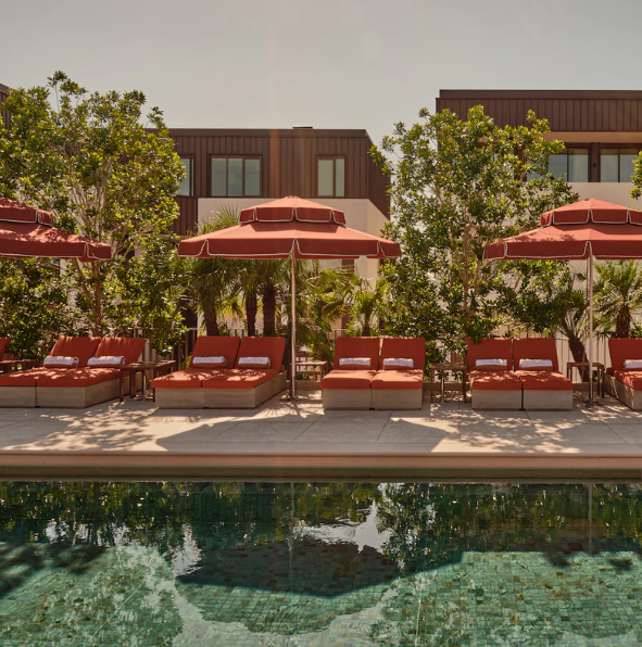 Swimming pool with lounge chairs and red umbrellas, surrounded by greenery and modern buildings in the background.