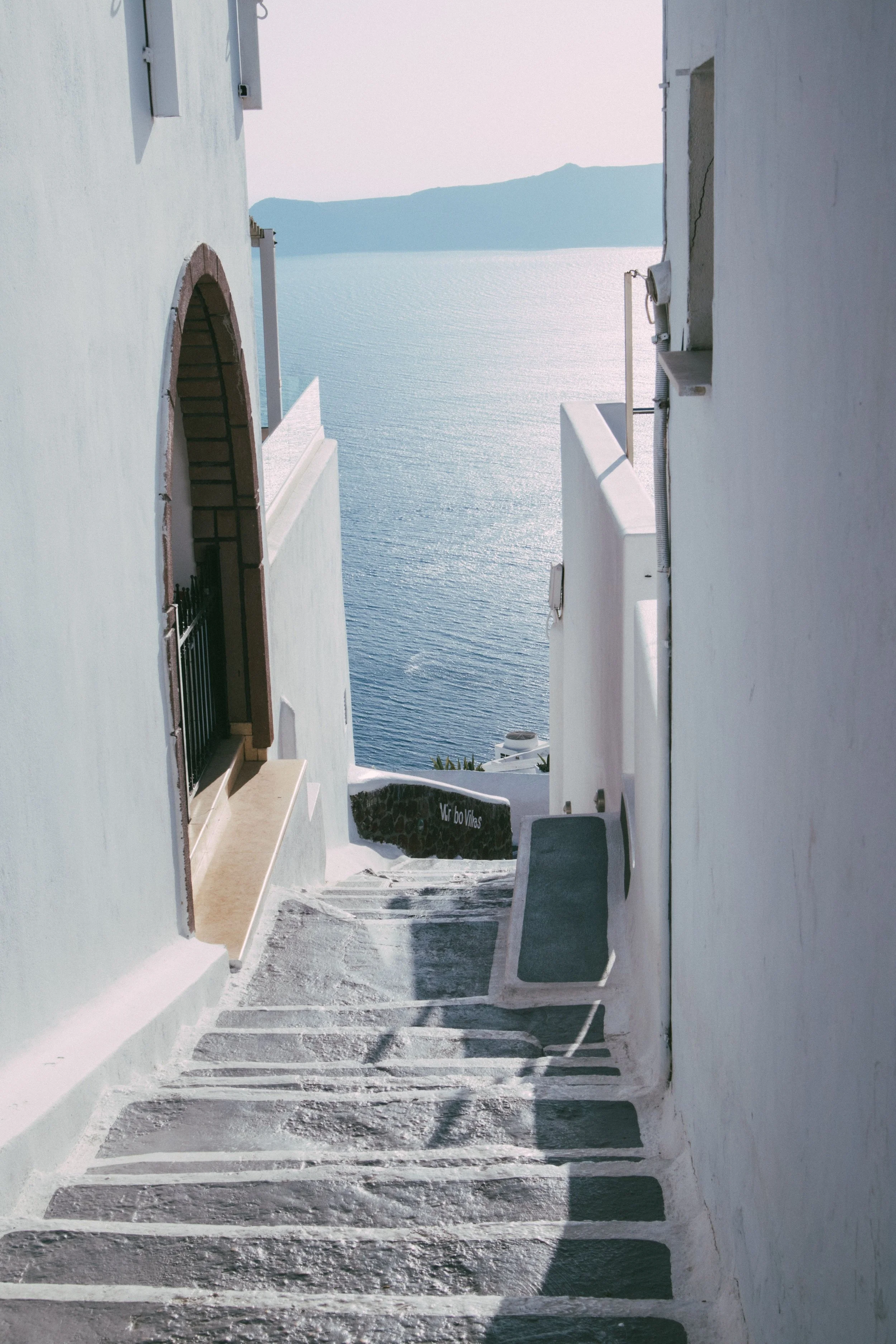 White-washed stairs descending between white buildings towards the sea, with a view of distant mountains and calm water.