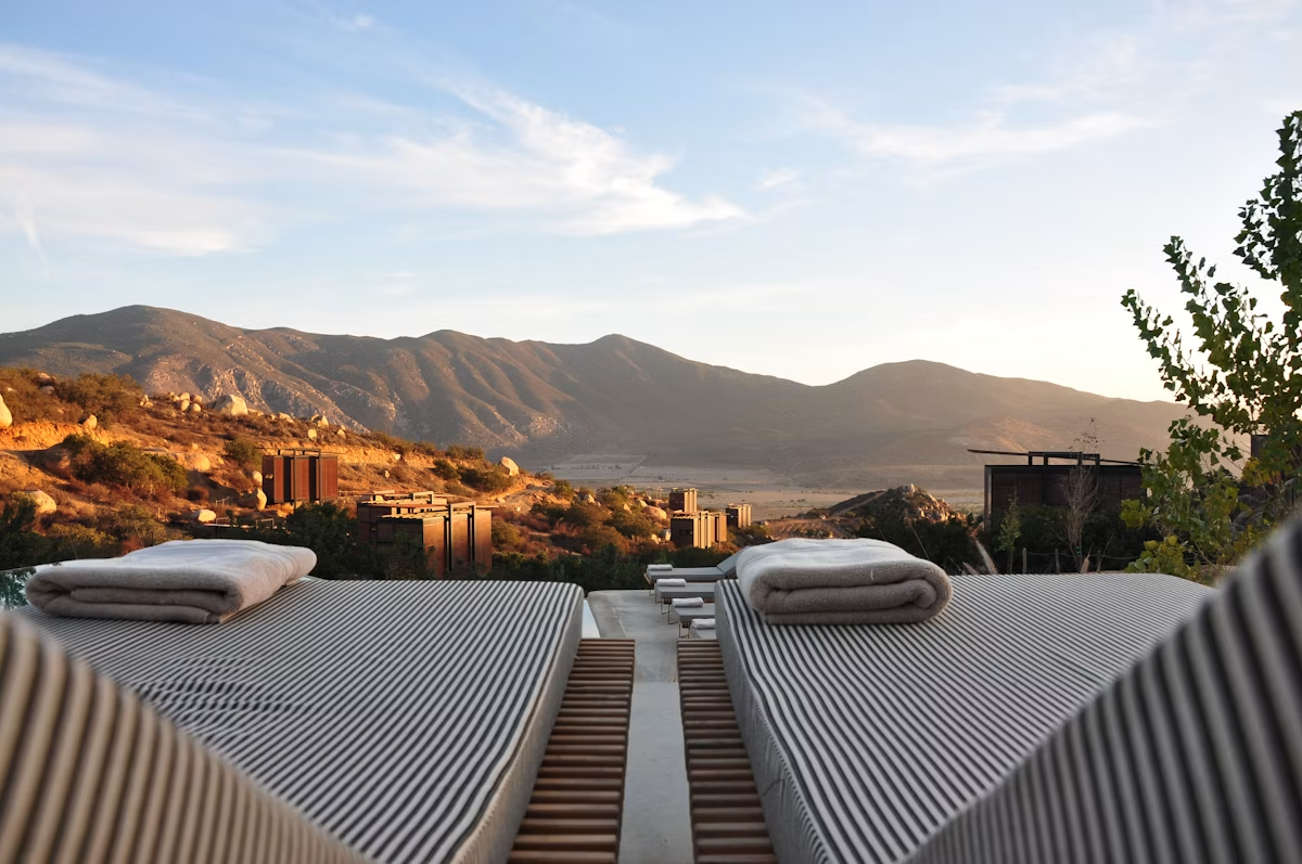 Two outdoor lounge chairs with rolled towels on top, overlooking a mountainous landscape at sunset with modern buildings in the distance.