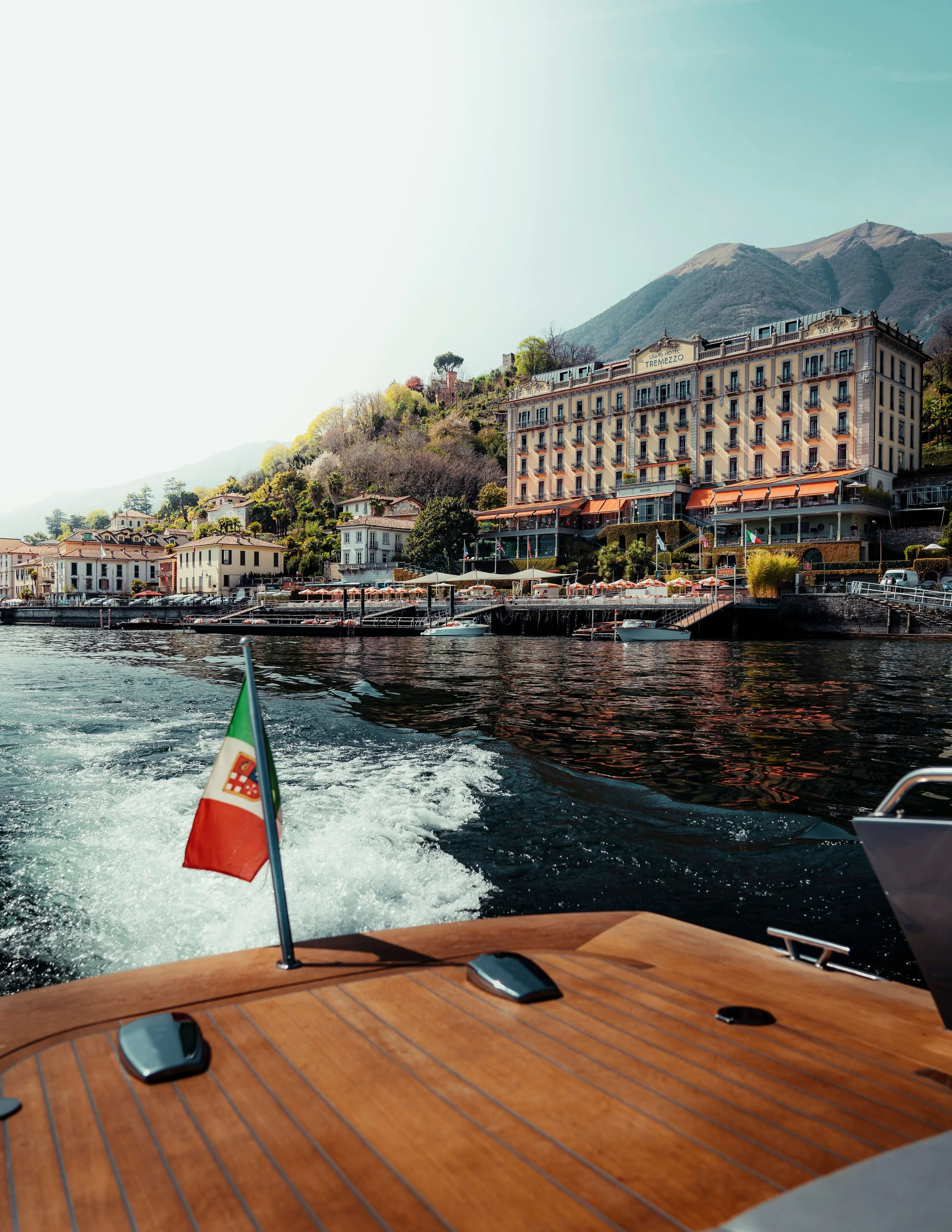 View from a boat showing a wooden deck, an Italian flag, and the water, with city buildings and mountains in the background.