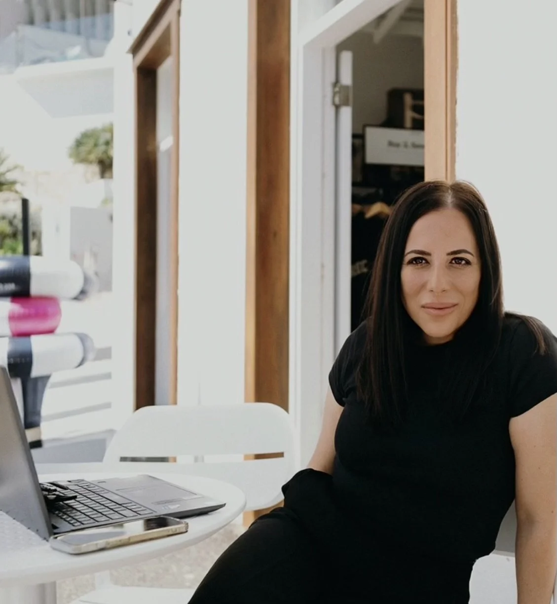 A woman with straight black hair sitting at a white table in front of a laptop and phone, with bookshelf and interior in the background.