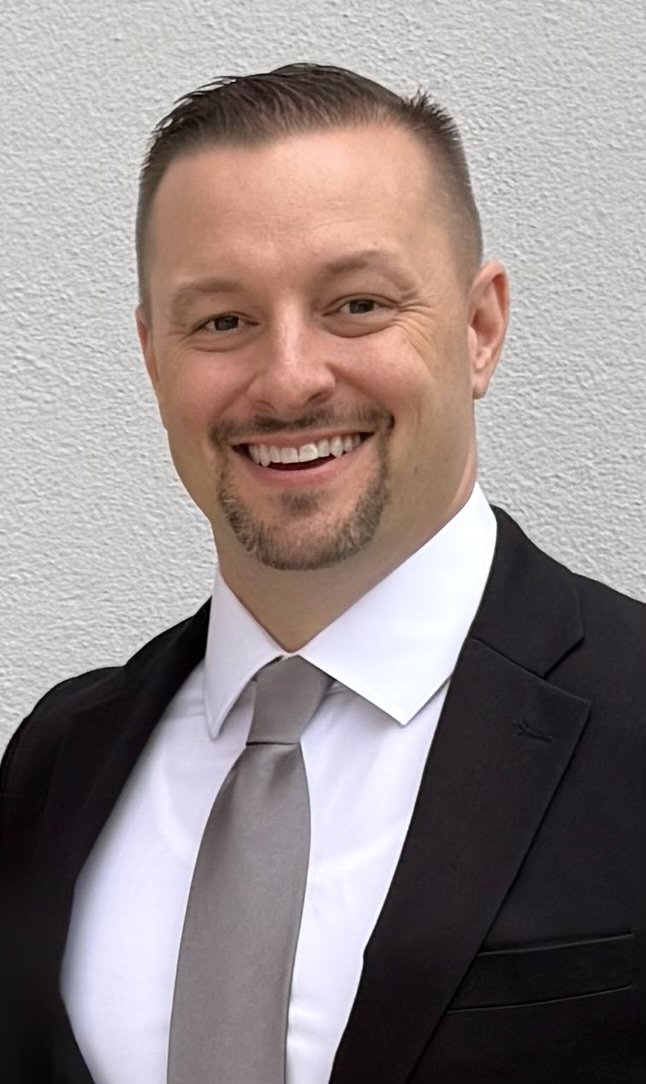A man with short brown hair and a goatee, dressed in a black suit, white shirt, and gray tie, smiling against a plain white textured background.