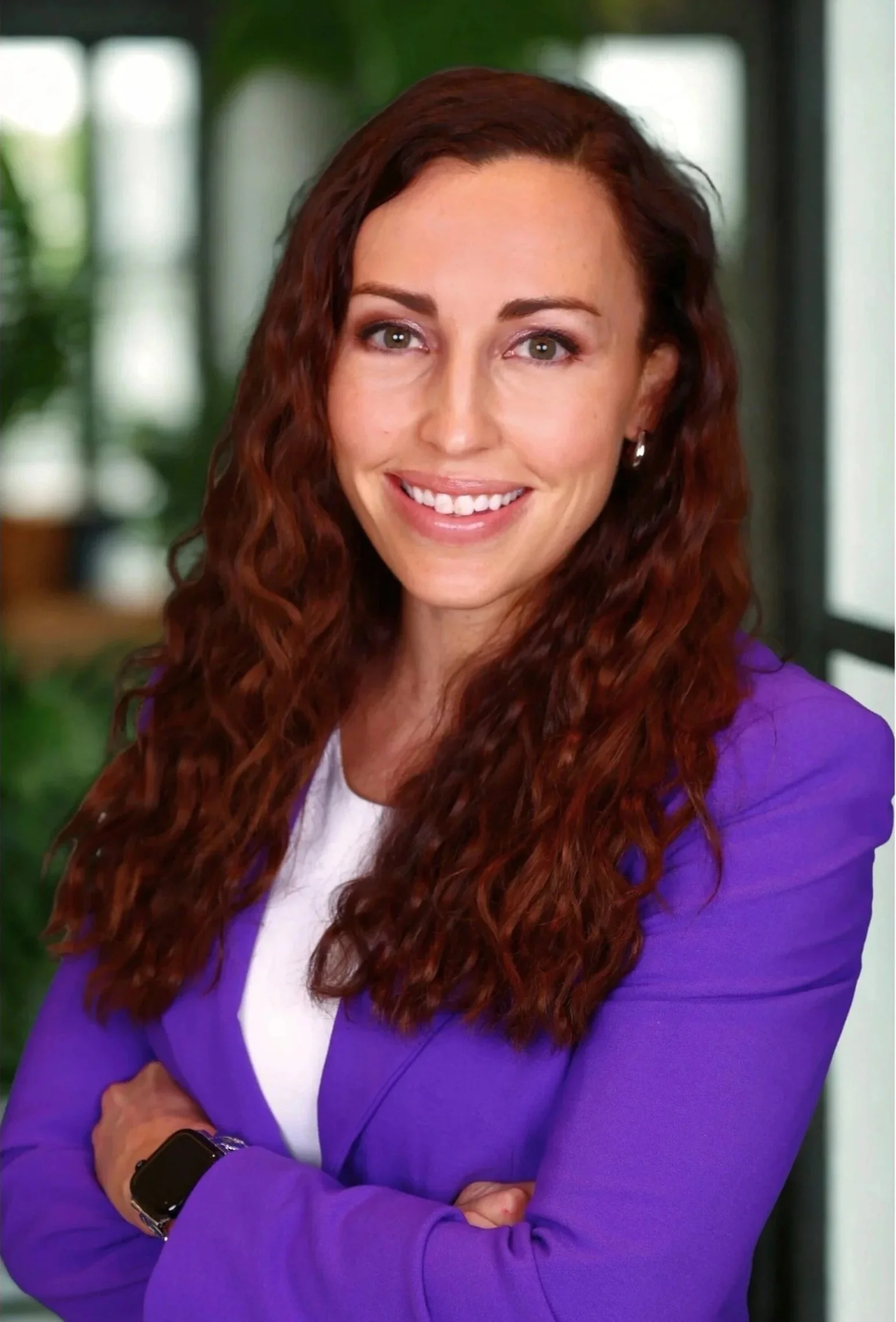 Woman with long, curly, red hair smiling, wearing a purple blazer and a smartwatch, standing indoors with greenery in the background.