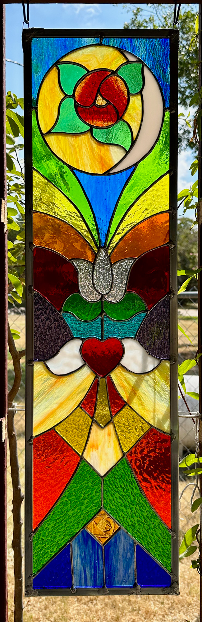Colorful stained glass window with abstract floral and natural shapes, including a red rose at the top, a white butterfly in the middle, a red heart below it, and various shapes and colors resembling flowers and leaves.