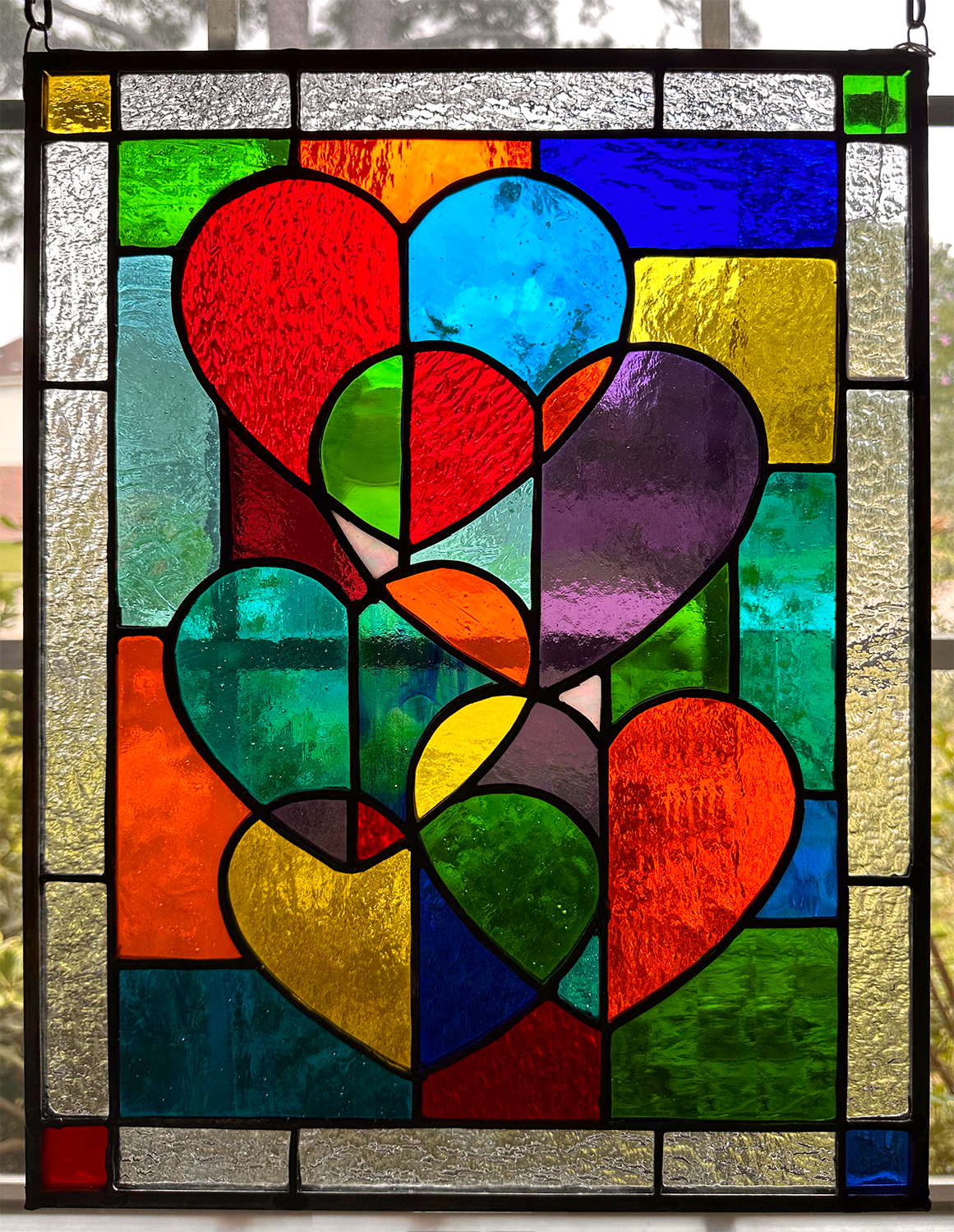 Colorful stained glass window featuring multiple hearts outlined in black, with various vibrant colors including red, blue, yellow, purple, green, and orange, set against a window with a blurred outdoor background.