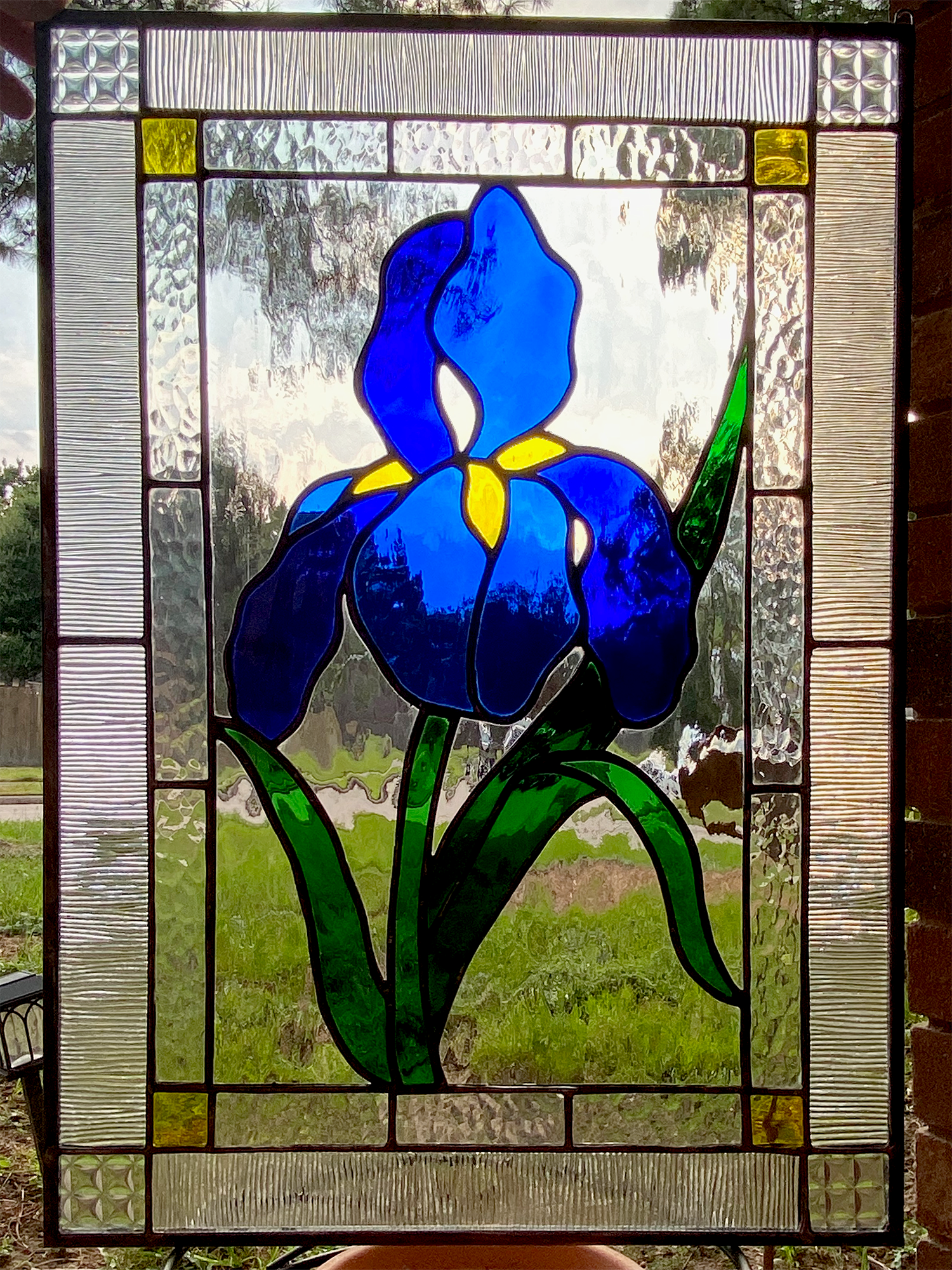 A stained glass window featuring a stylized blue iris flower with green leaves and a yellow center, framed by rectangular glass pieces with textured and clear patterns.