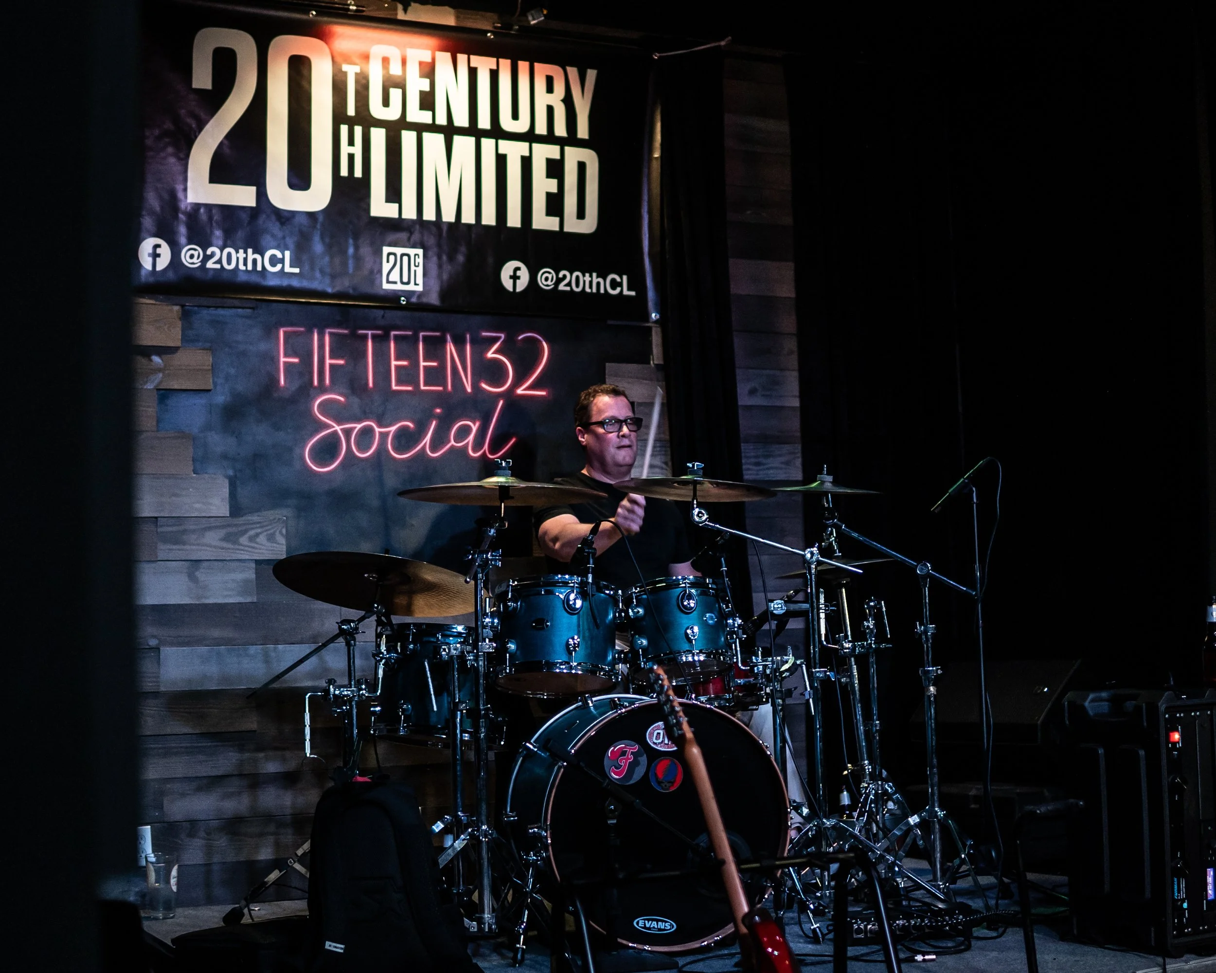 A man playing the drums on a stage at a live music venue, with signs behind him reading '20th Century Limited' and 'Fifteen32 Social'.