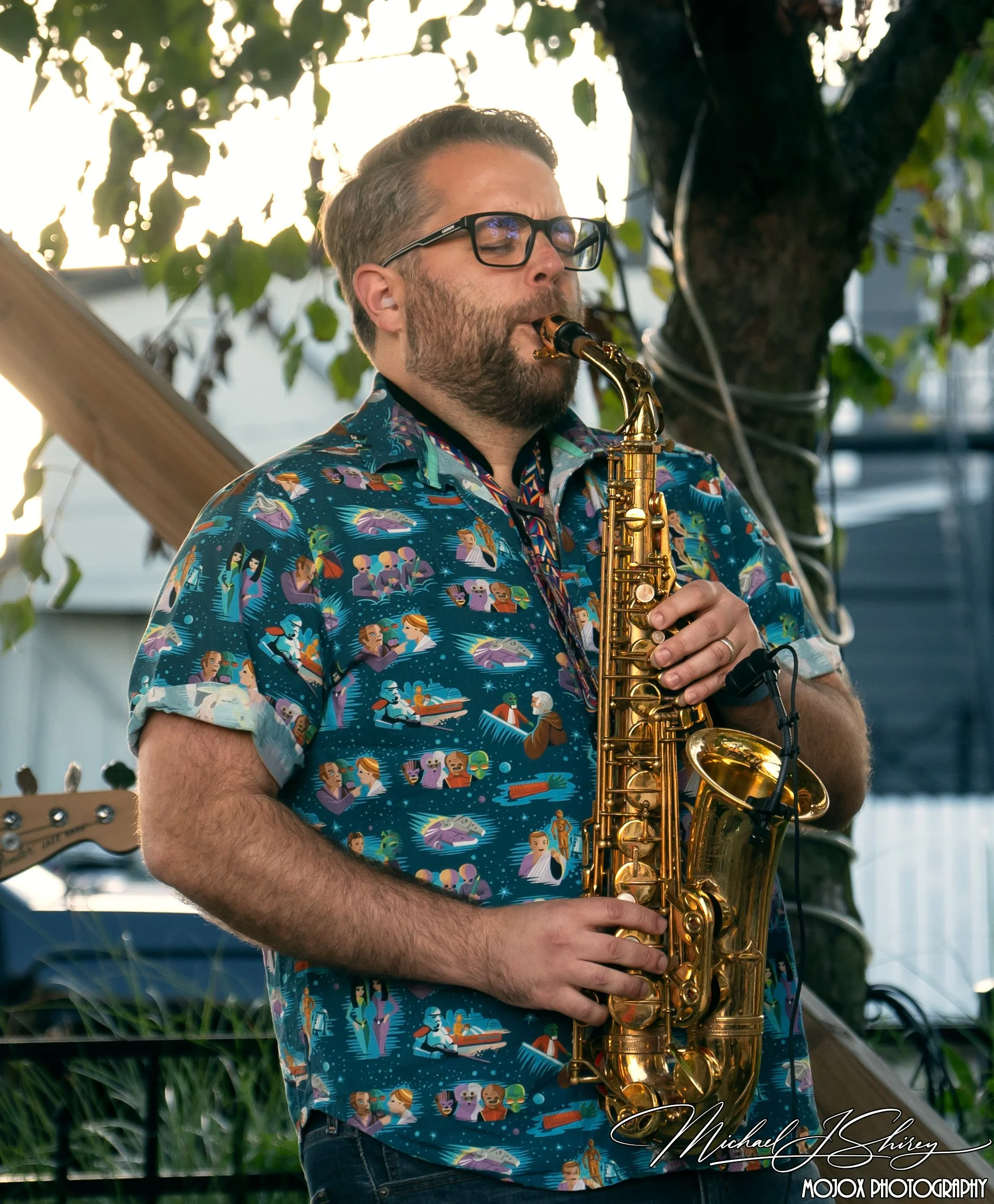 A man playing a gold saxophone outdoors, wearing glasses and a colorful Hawaiian shirt with various animated characters.