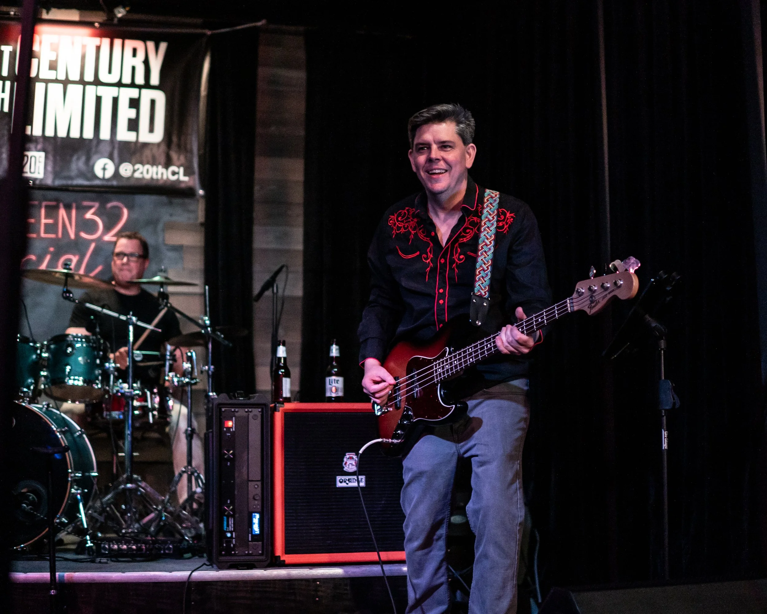Two musicians perform on stage, one playing drums and the other playing a bass guitar, with a banner in the background.