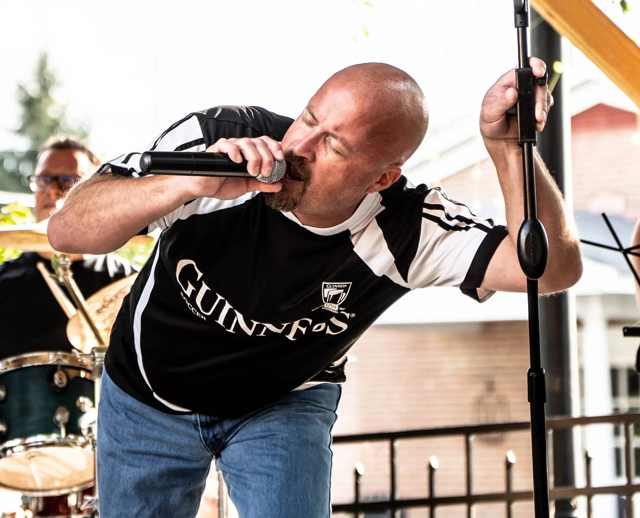 A man singing into a microphone on an outdoor stage, wearing a black and white sports jersey with the Guinness logo.