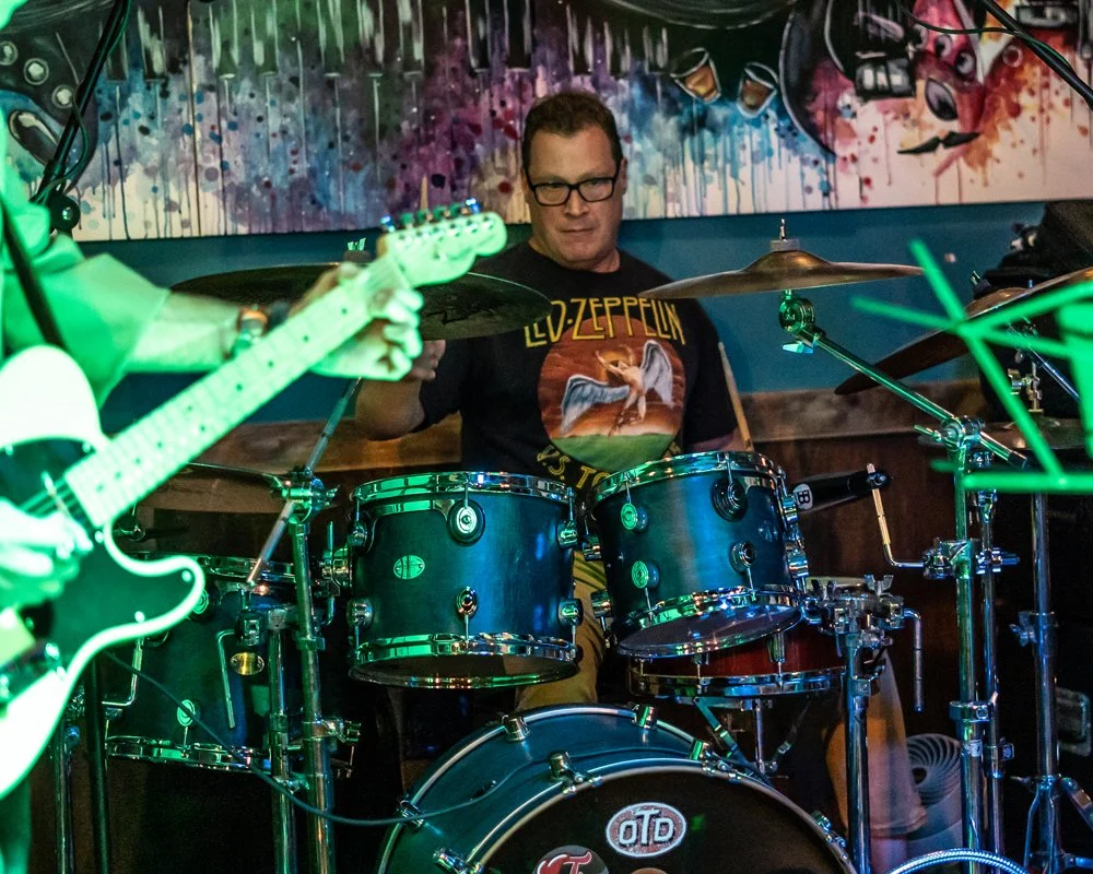 A man wearing glasses and a Led Zeppelin t-shirt playing drums during a live music performance, with a guitarist visible on the left, in a dimly lit venue with colorful artwork on the wall behind them.