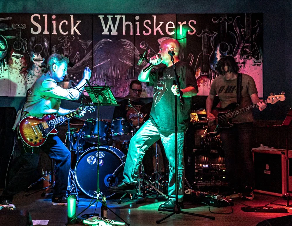 A four-member band performing live on stage with a colorful mural backdrop reading 'Slick Whiskers', featuring a person playing electric guitar, a drummer, a vocalist holding a microphone, and a bassist with an electric bass guitar.