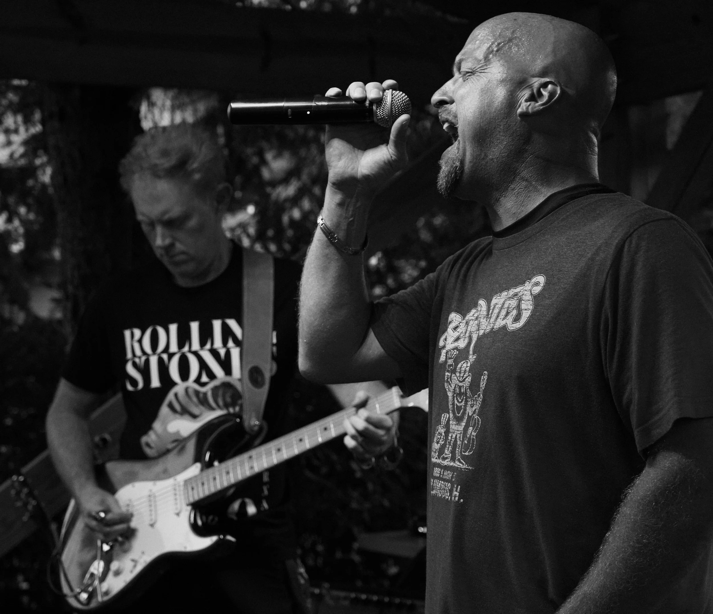 A man singing into a microphone and a man playing guitar during an outdoor performance, black and white photo.