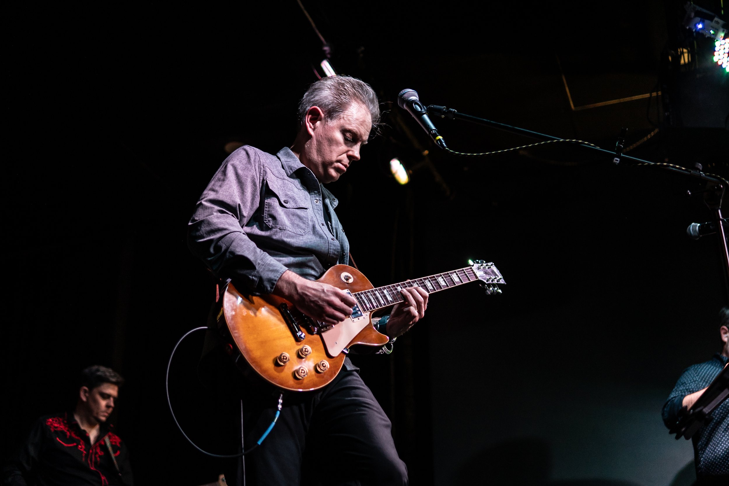 A musician playing a brown electric guitar on stage, with two other band members in the background, under stage lighting.