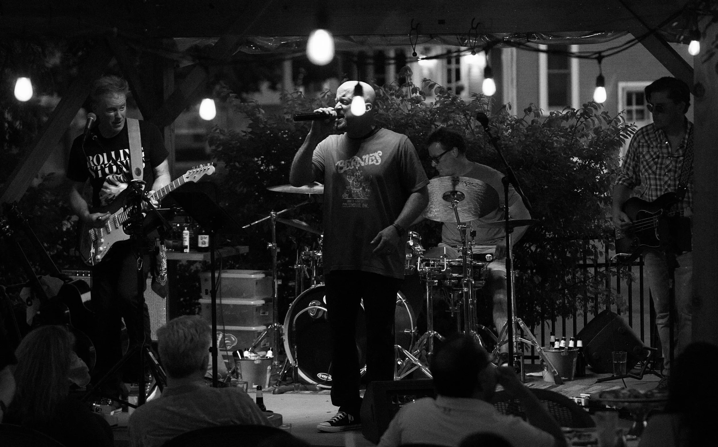 A band performing live on an outdoor stage with string lights overhead, featuring a singer, guitarist, drummer, and bassist.