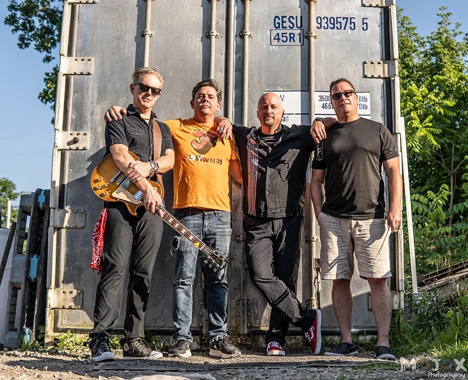 Four men standing together outdoors in front of a large metal container. One man holds a guitar, and they are all casually dressed, smiling or with relaxed expressions.