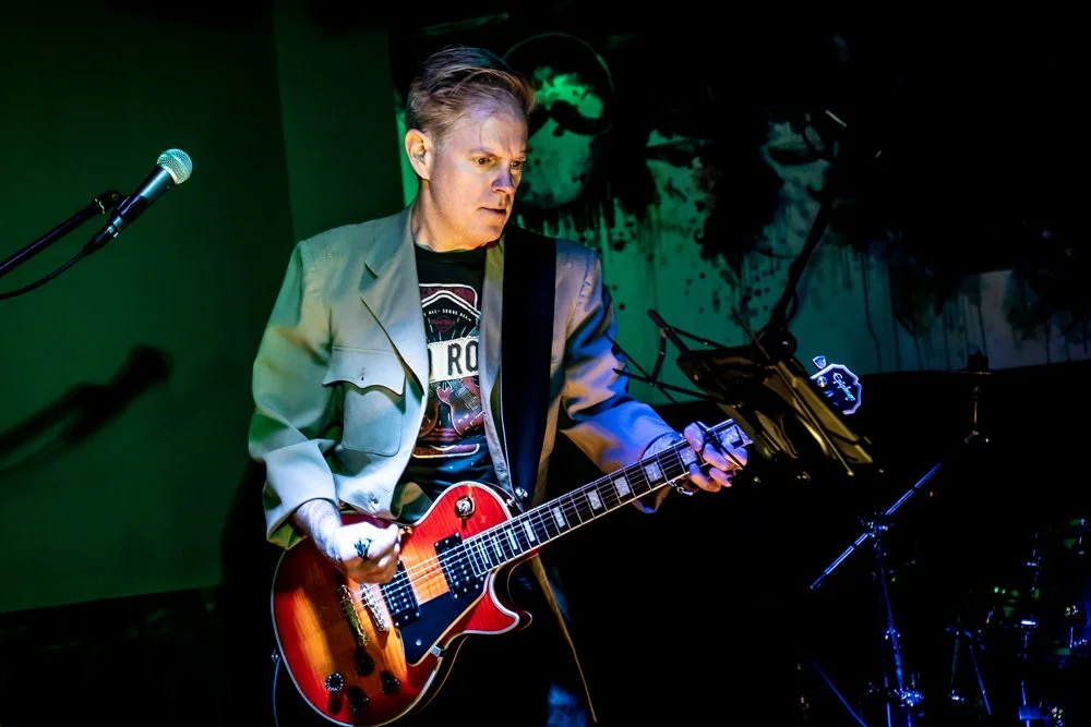 A musician playing an electric guitar on stage, wearing a beige jacket and a black graphic t-shirt, with a microphone stand nearby and a dark, artistic backdrop.