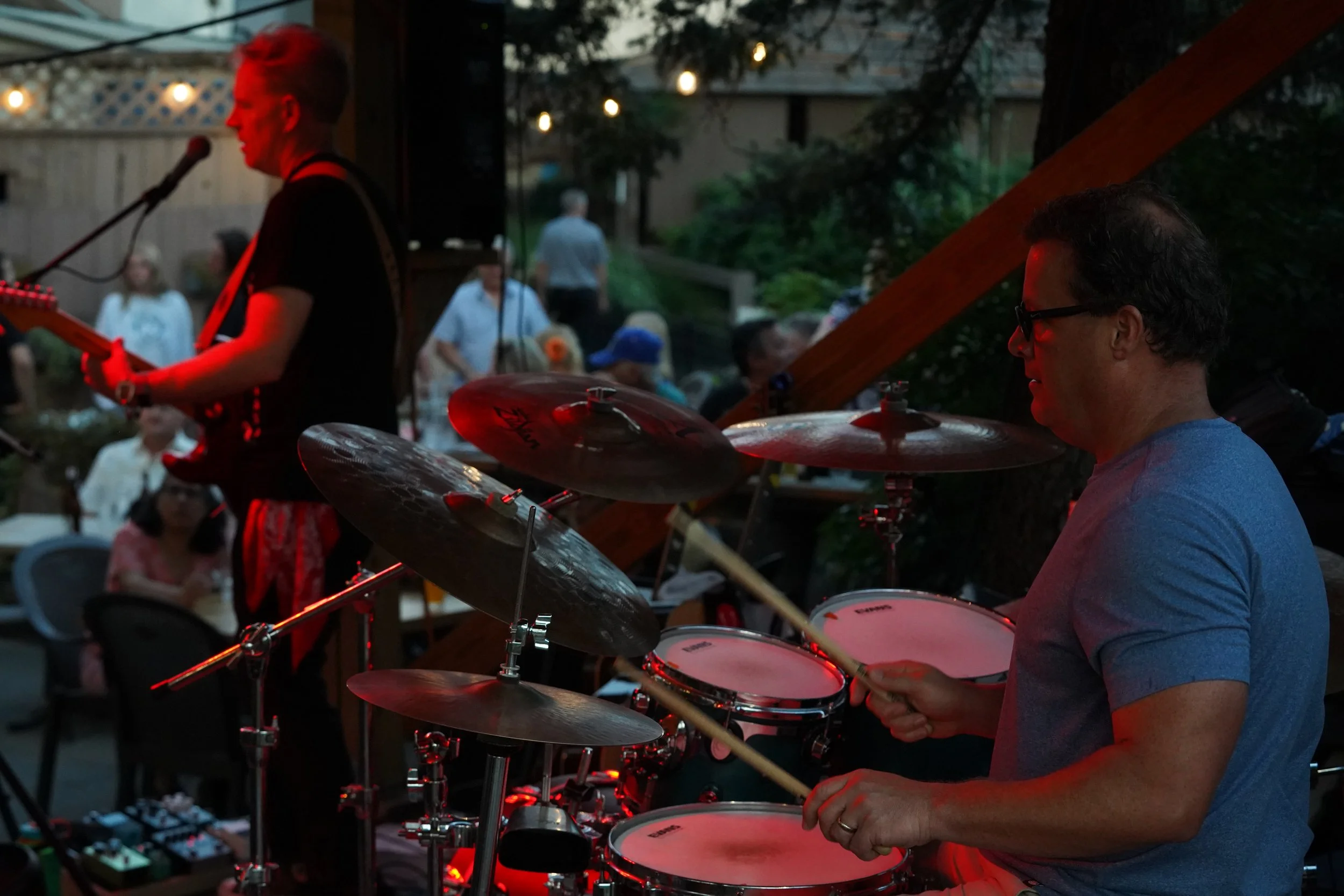 A man playing drums at an outdoor concert during evening, with a guitarist performing on stage in the background, and audience members seated in the background