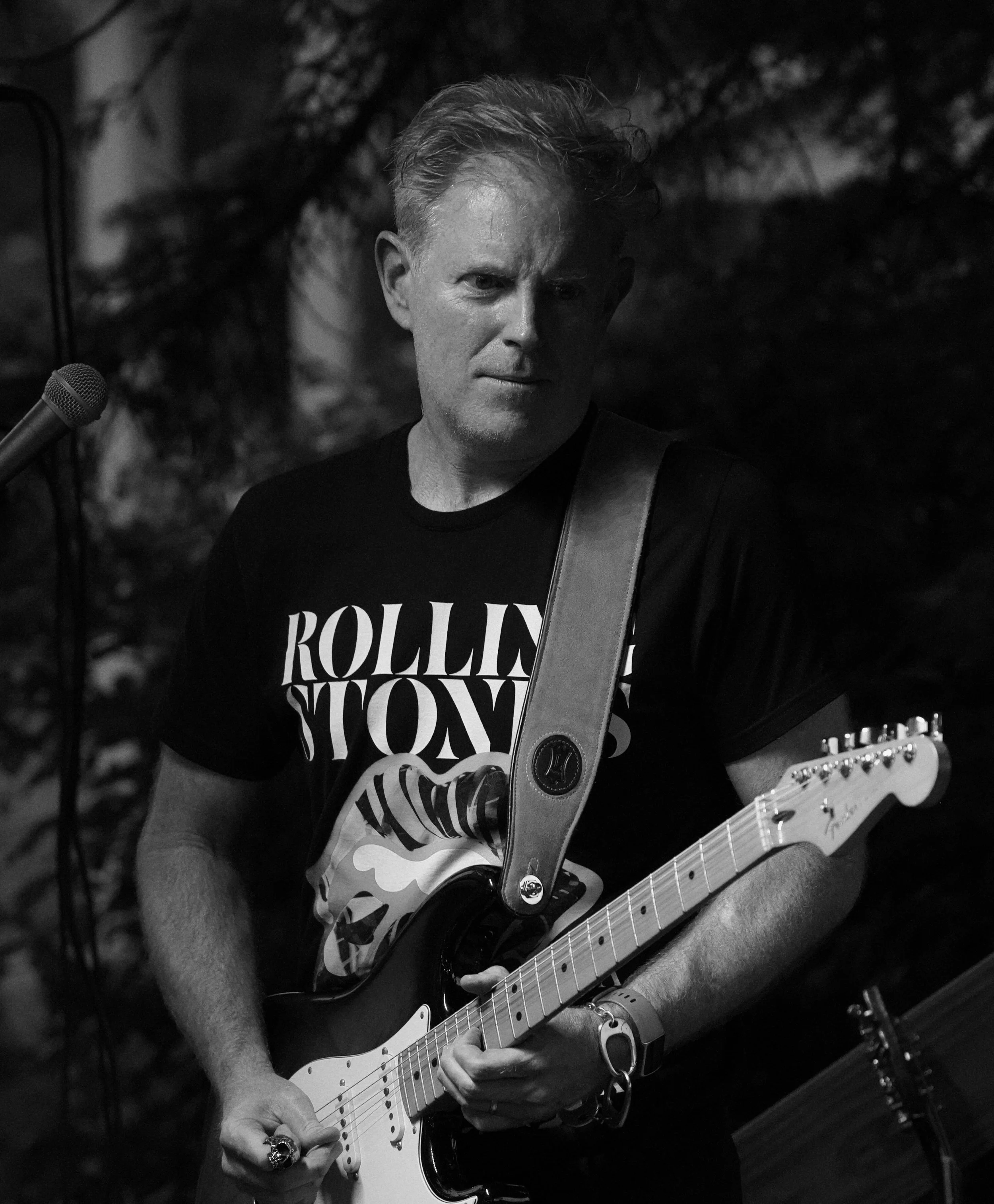 Black and white photo of a man playing an electric guitar, wearing a t-shirt with 'Rolling Stones' lettering, outdoor setting with trees, and a microphone in the background.
