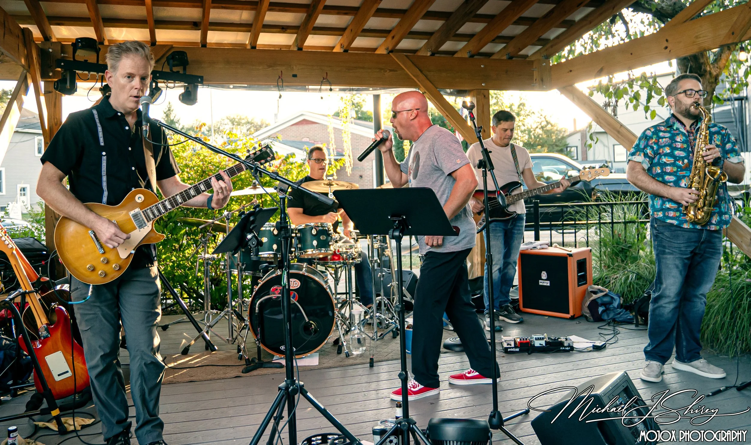 A band performing live on an outdoor stage with a wooden roof, featuring a guitarist, a vocalist, a drummer, a bassist, and a saxophonist, with various musical equipment around.