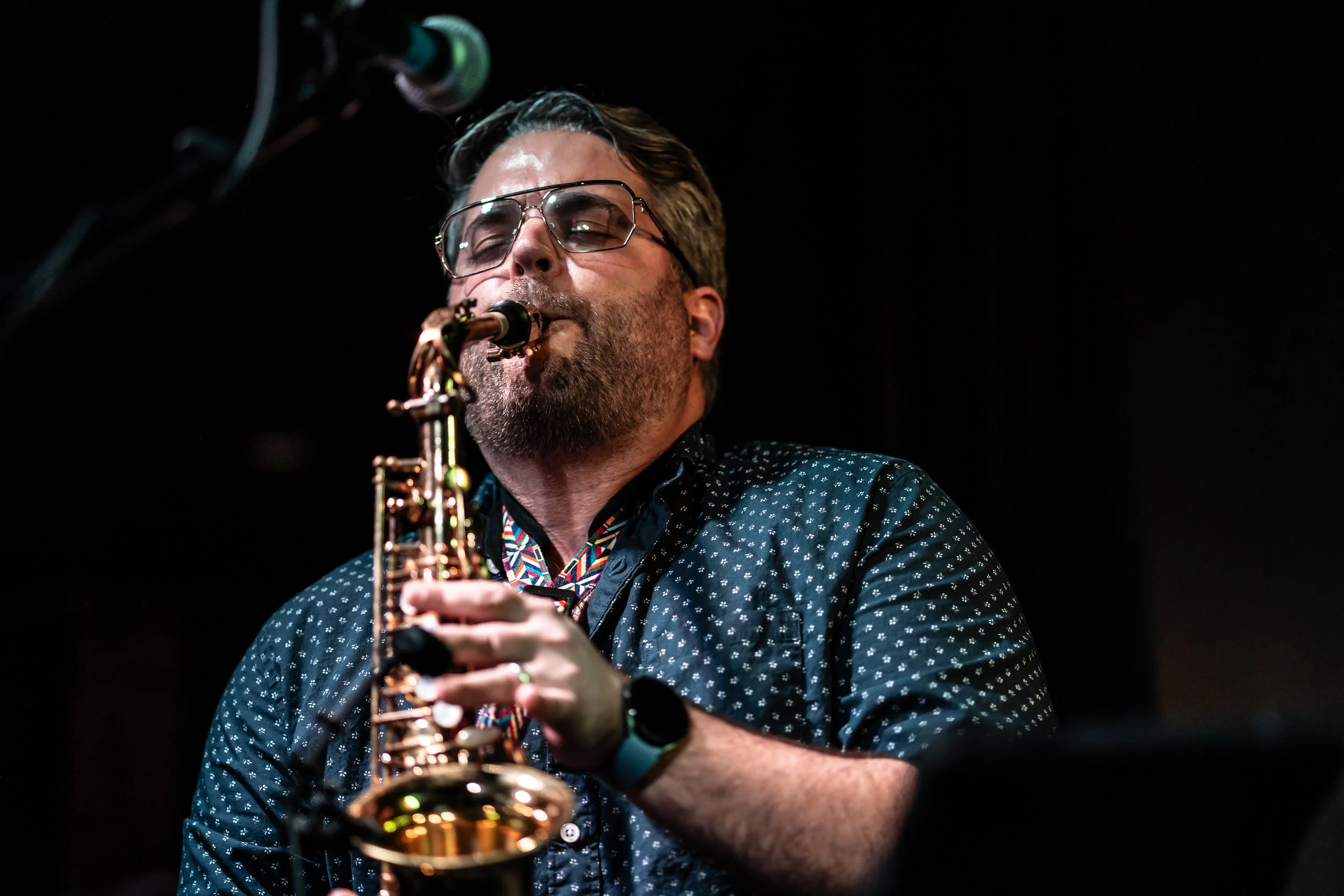 A man playing a saxophone on stage, wearing glasses, a patterned dark shirt, and a watch, with a microphone nearby.